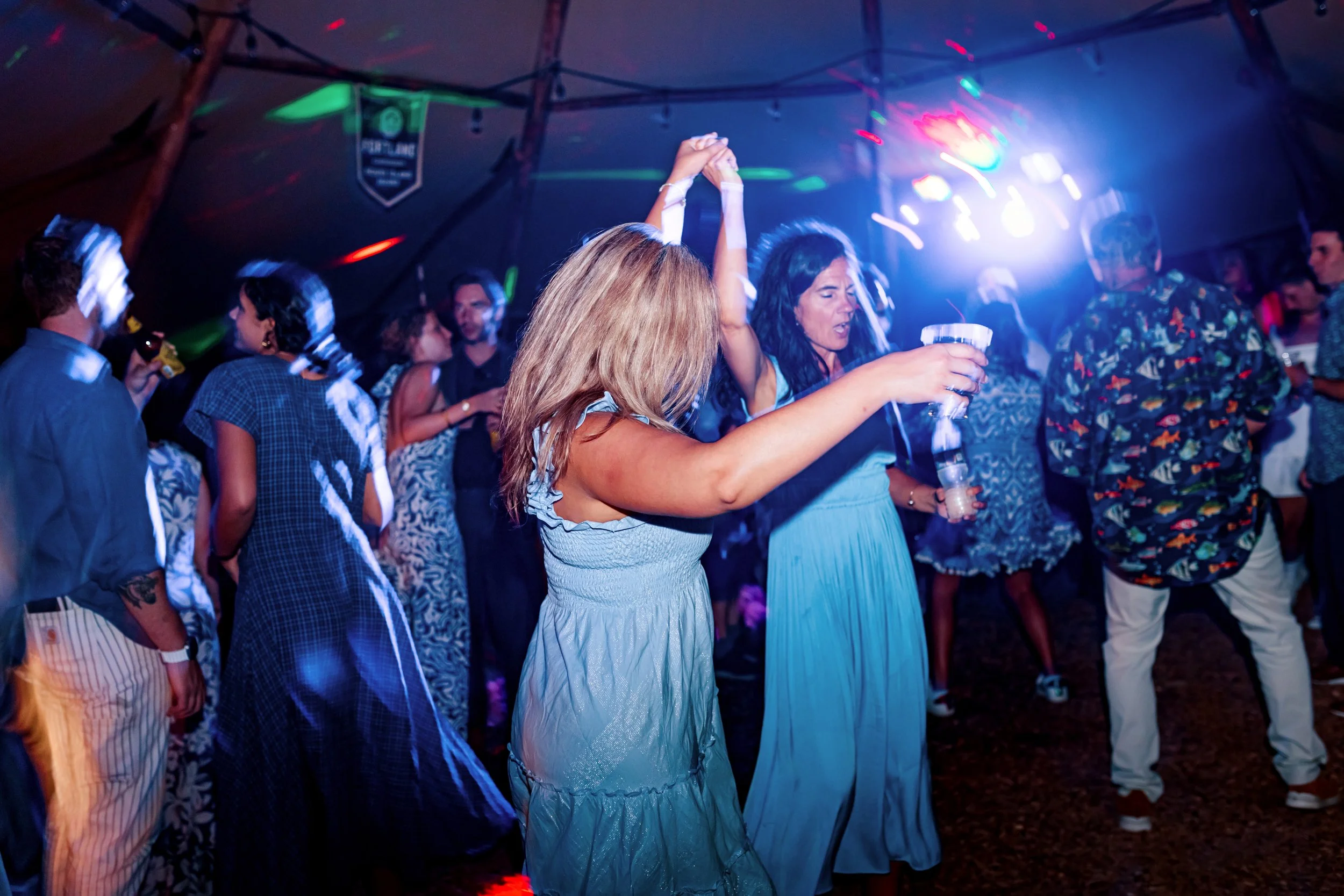 People dancing and enjoying a party under colorful lights at a tented event.