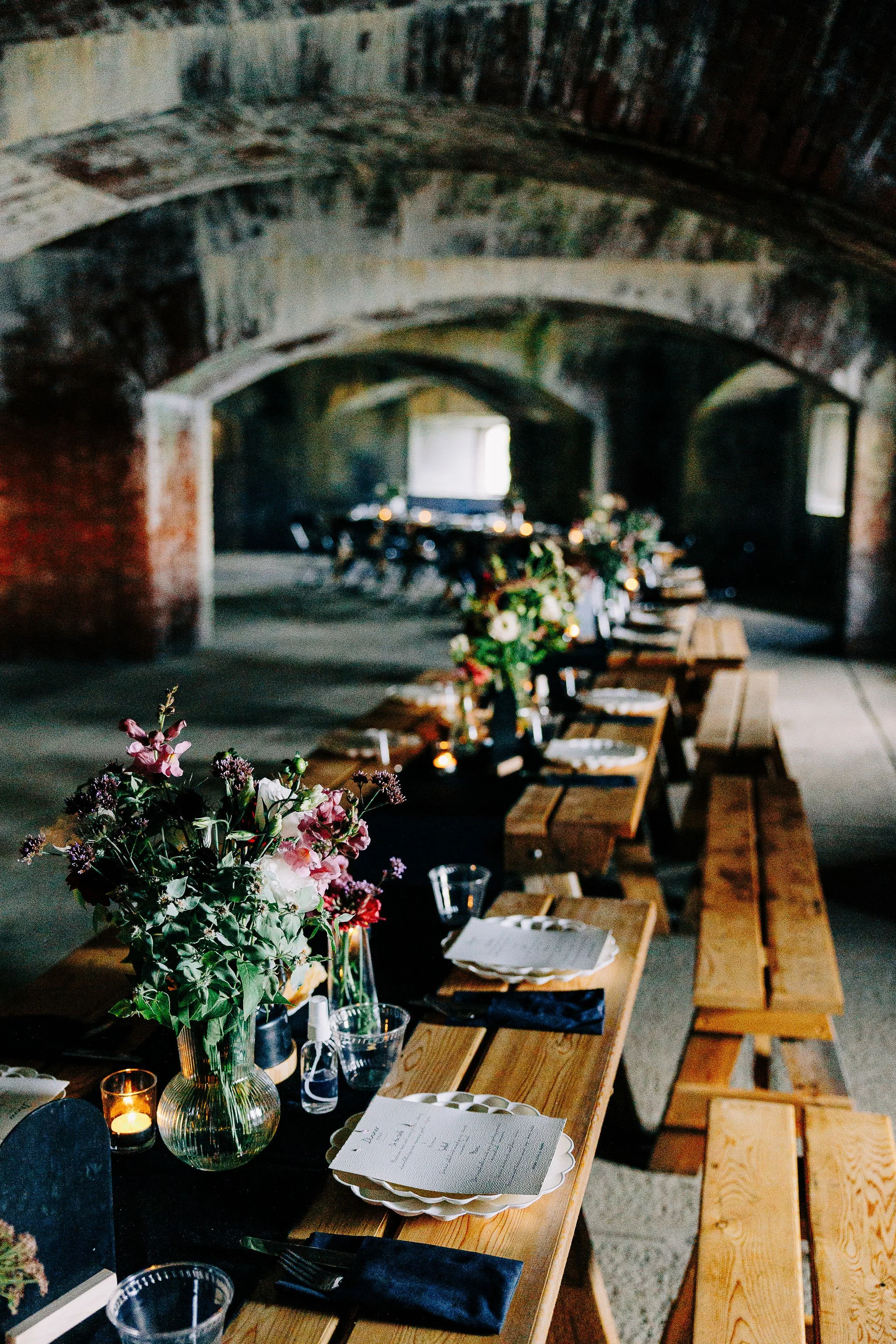 Long rustic wooden banquet table set with floral centerpieces, candles, plates, glasses, and napkins inside an abandoned brick tunnel with archways.