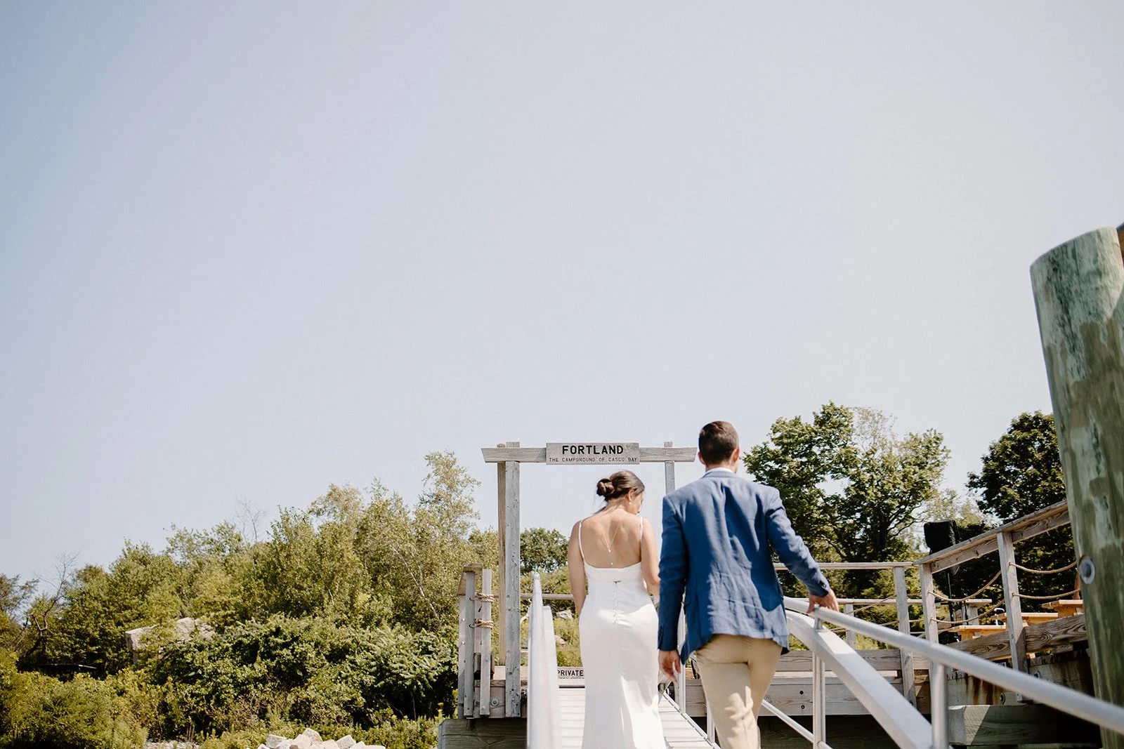 A bride in a white wedding dress and a groom in a blue blazer walk up a wooden ramp toward a sign that reads "Fortland, the campground of Casco Bay," surrounded by green trees on a clear day.