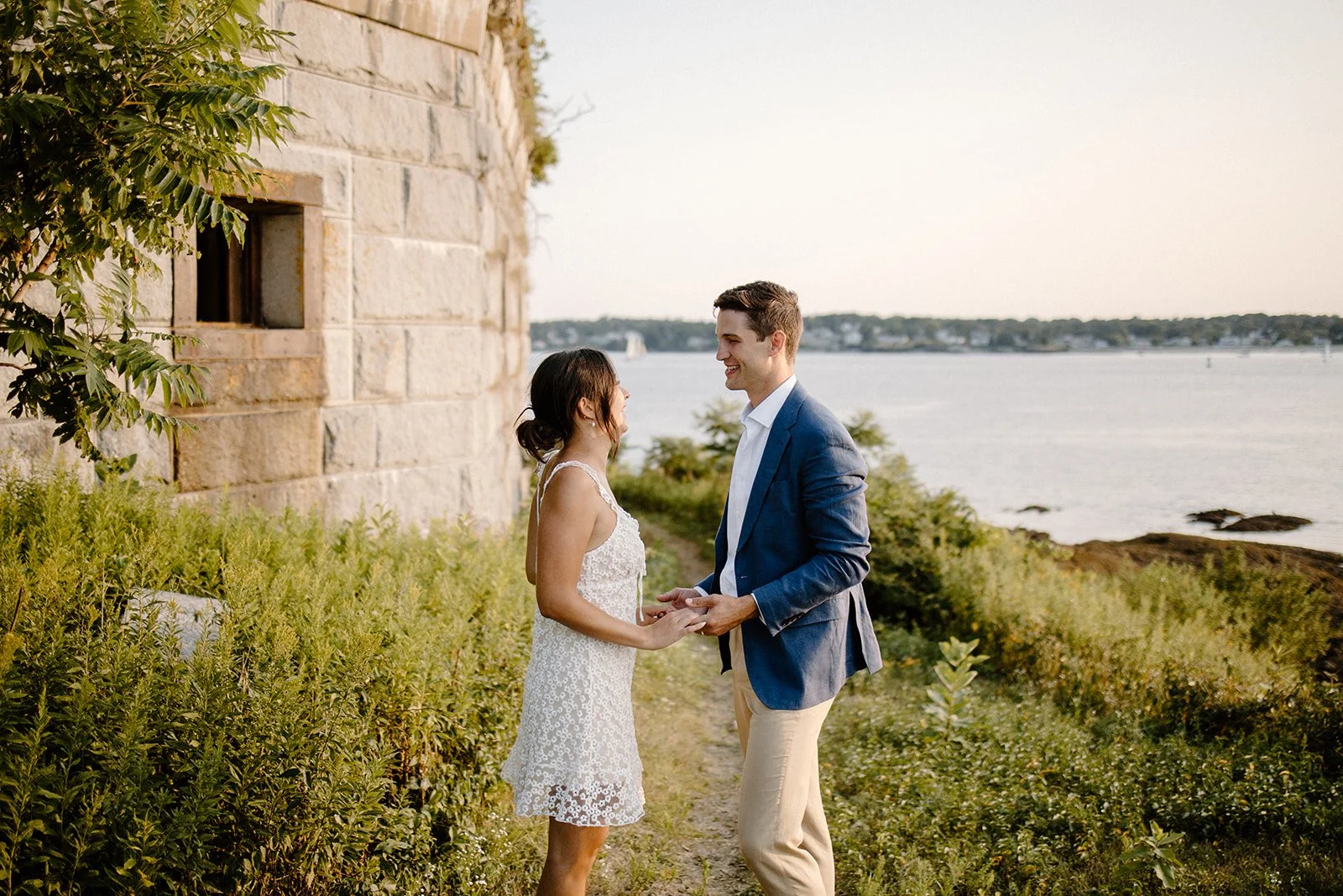 A couple stands outside near a stone building, holding hands and smiling at each other, with water and a distant horizon in the background.