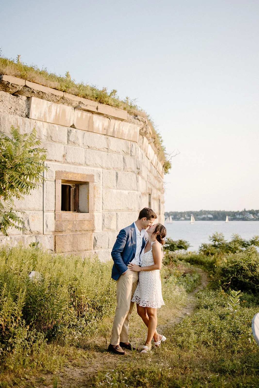 A couple is sharing a romantic moment by a river or lake, with sailboats in the background and a stone building wall to the left.