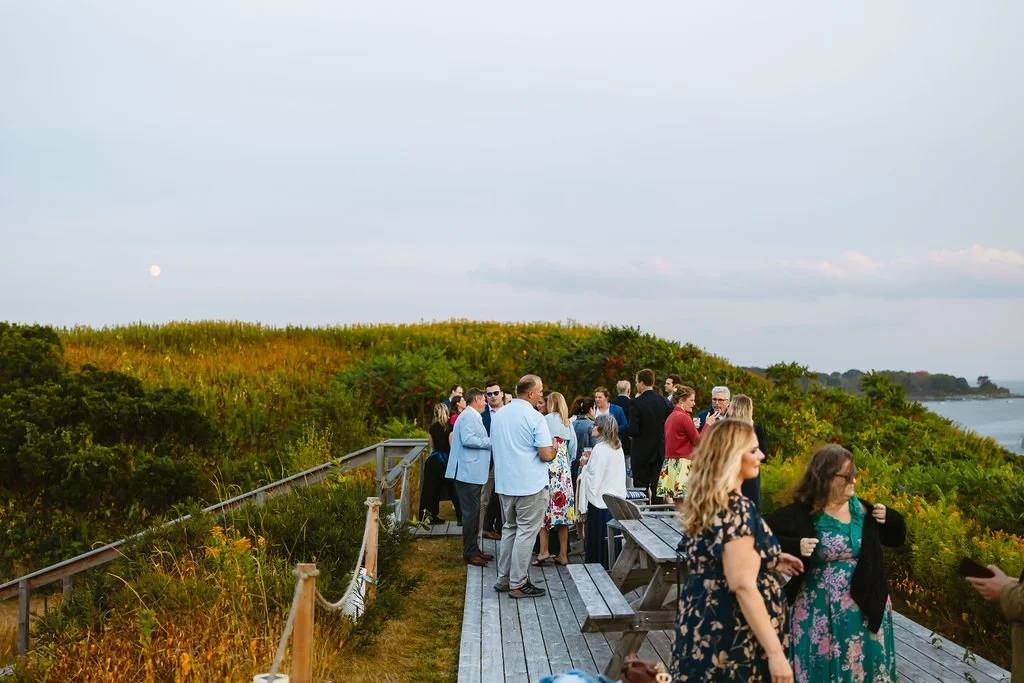 People gathered on a wooden deck with benches, overlooking a lush green hill and a body of water, during sunset or early evening.