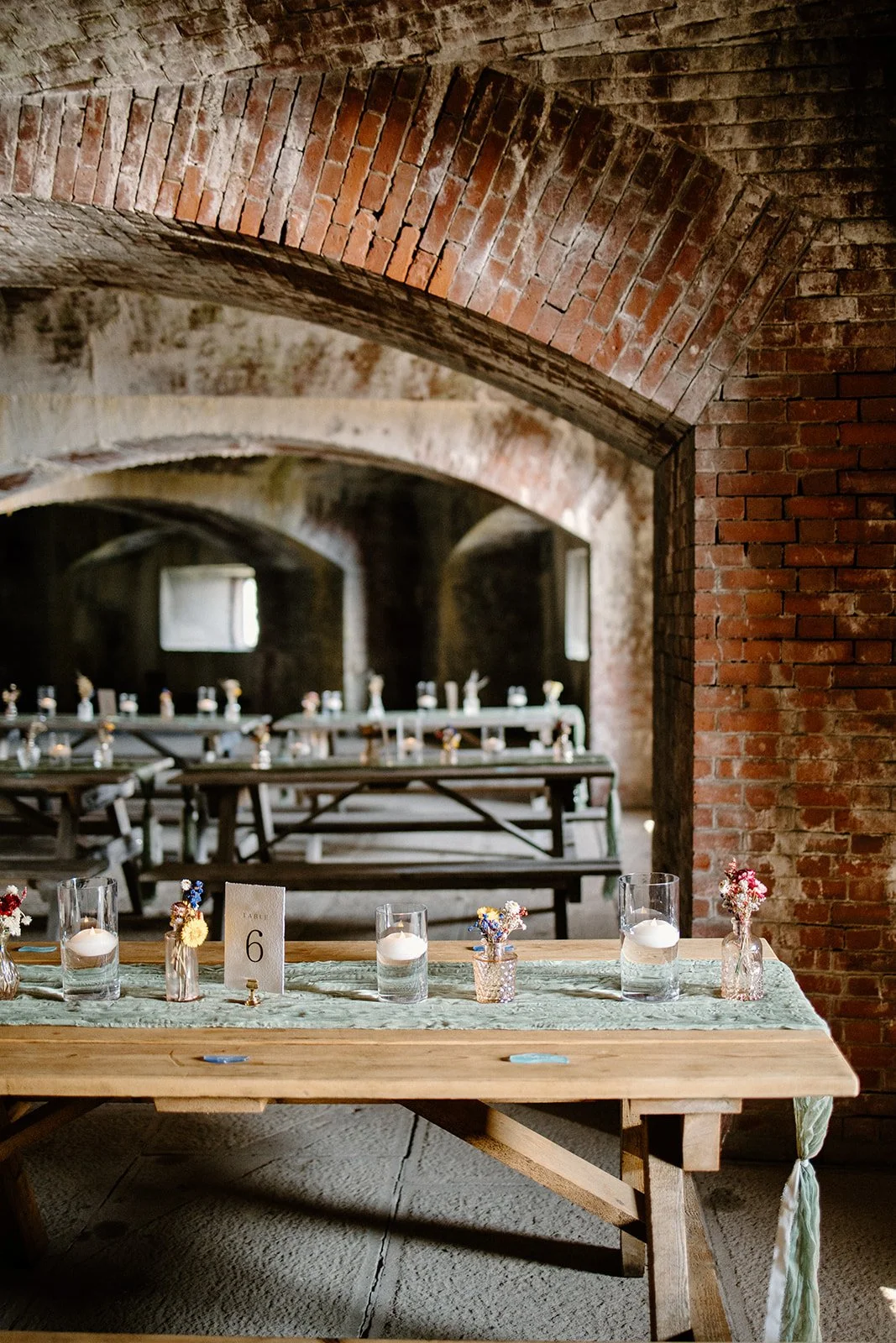 A rustic event space featuring brick arch ceilings, with a long wooden table decorated with vases of small flowers and candles, set for a gathering.