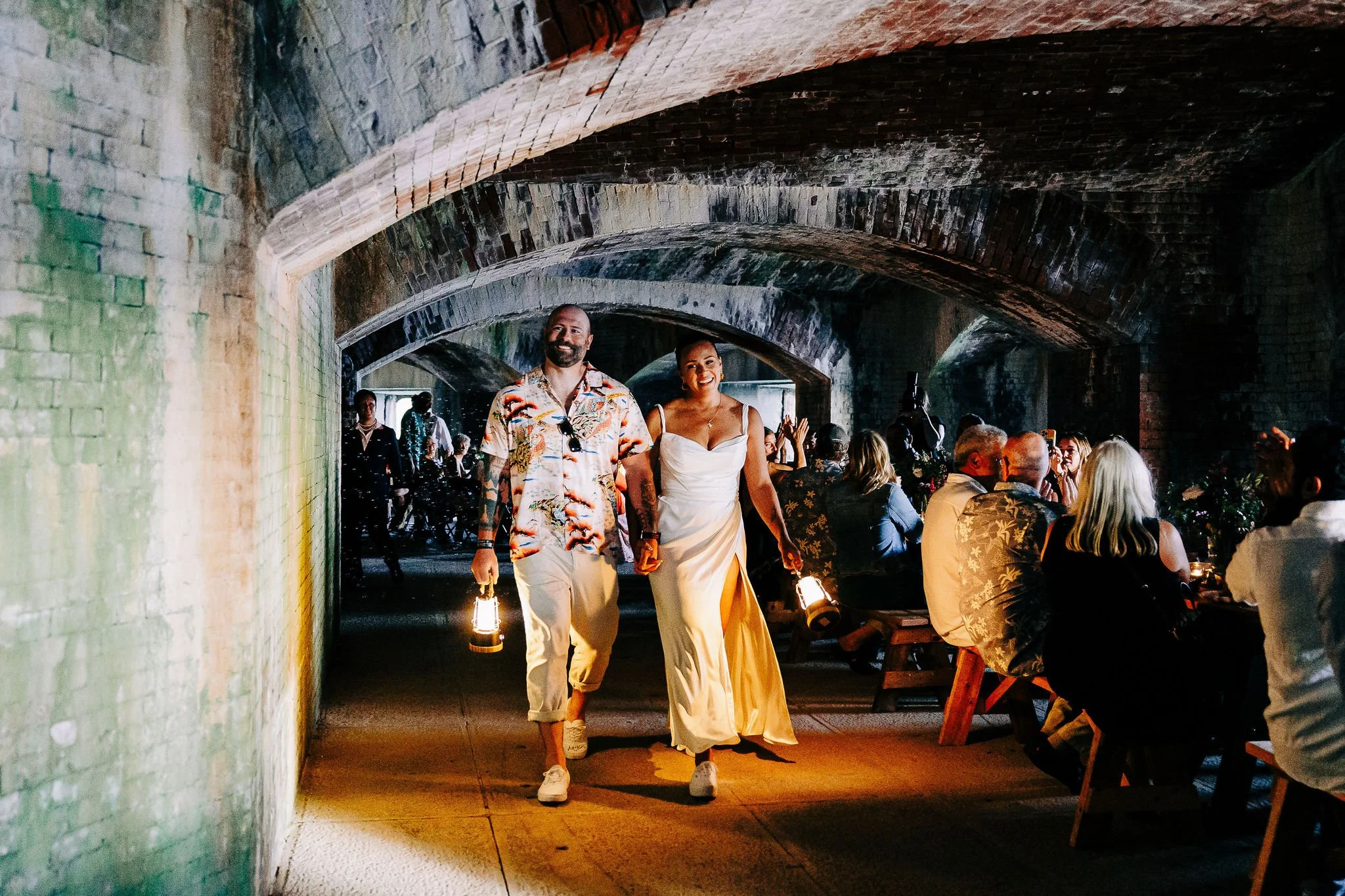 A couple walking through a tunnel at a lively event, with other guests seated at tables in the background, illuminated by lanterns.