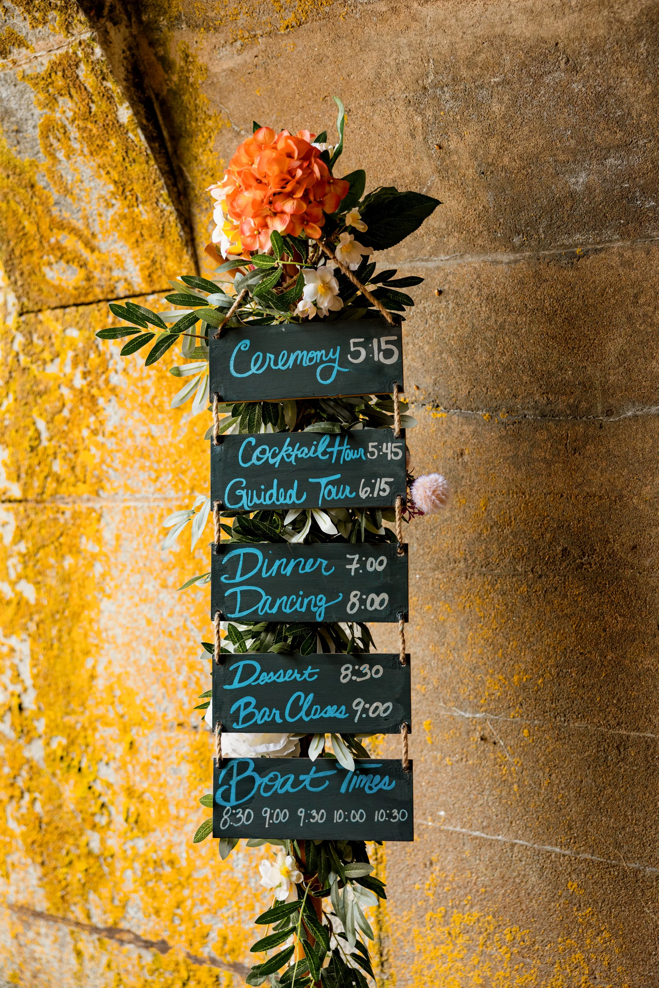 A decorative schedule sign with flowers and greenery on a stone wall, listing events and times for a wedding or celebration, including ceremony, cocktail hour, guided tour, dinner, dancing, dessert, bar closing, and boat times.