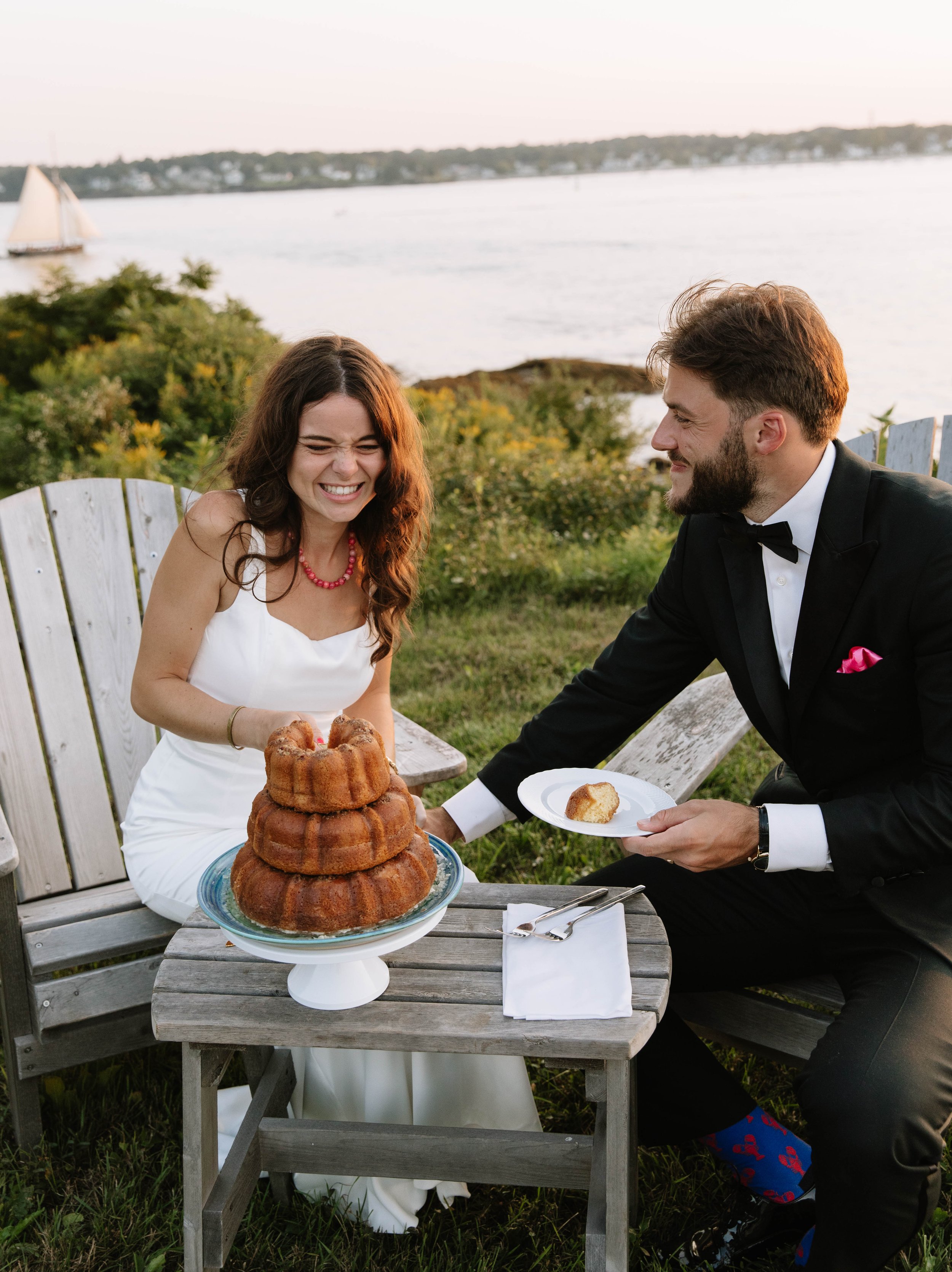 A couple in formal attire celebrating with cake outdoors by a river at sunset, with the woman smiling and the man holding a plate and a fork.