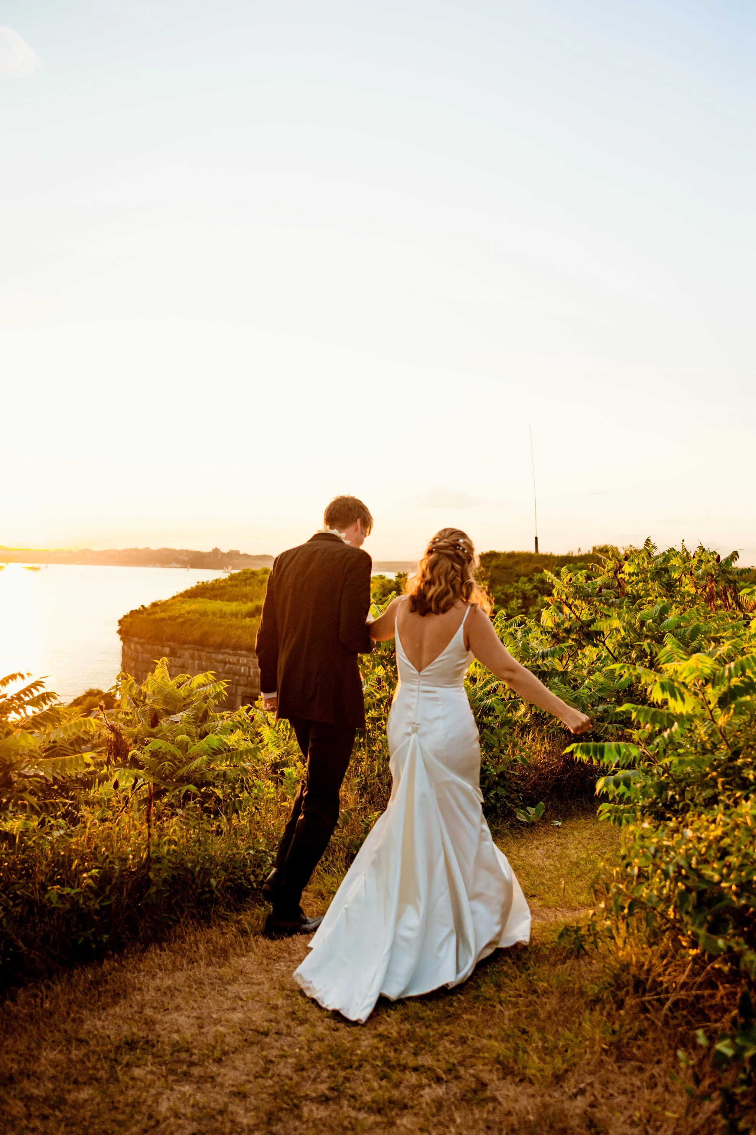 A couple dressed in wedding attire walking along a path on a grassy hill during sunset with water and trees in the background.