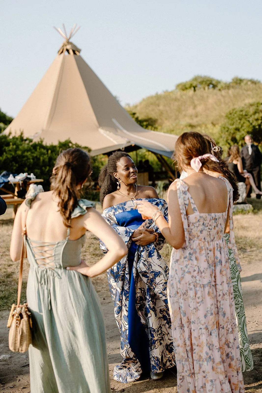 A group of women at an outdoor gathering, engaging in conversation near a large teepee-shaped tent in a grassy area, dressed in summer clothing.