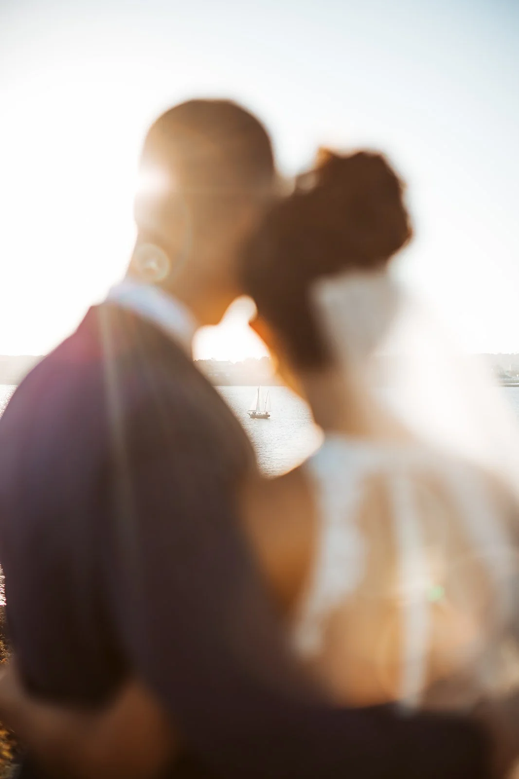 A blurry photo of a man and woman with their faces close together, backlit by the sun, with a body of water and sailboats in the background.