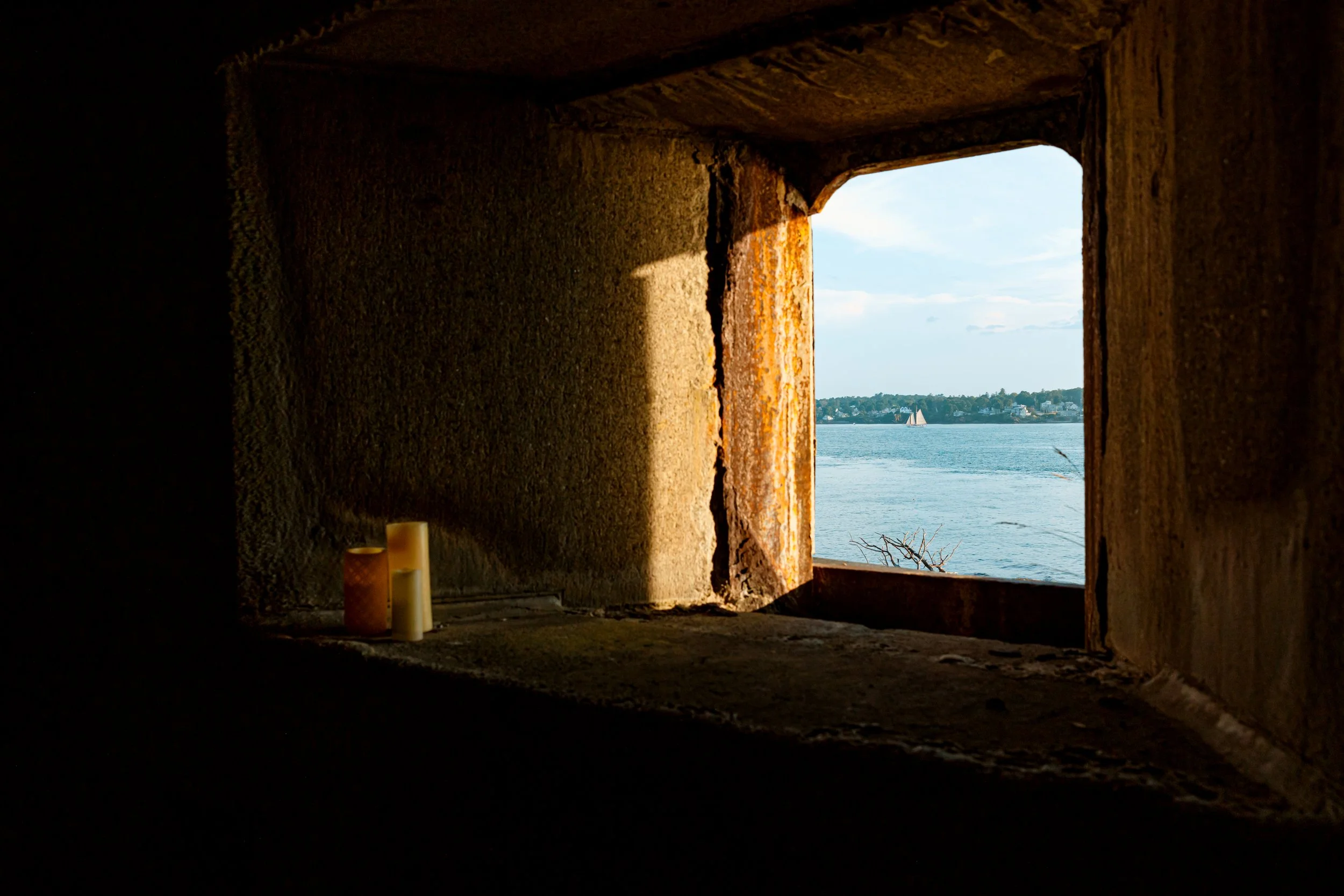 View of a river or large body of water through a small opening in an old stone or concrete structure, with candles on the left side, and a partly cloudy sky in the background.