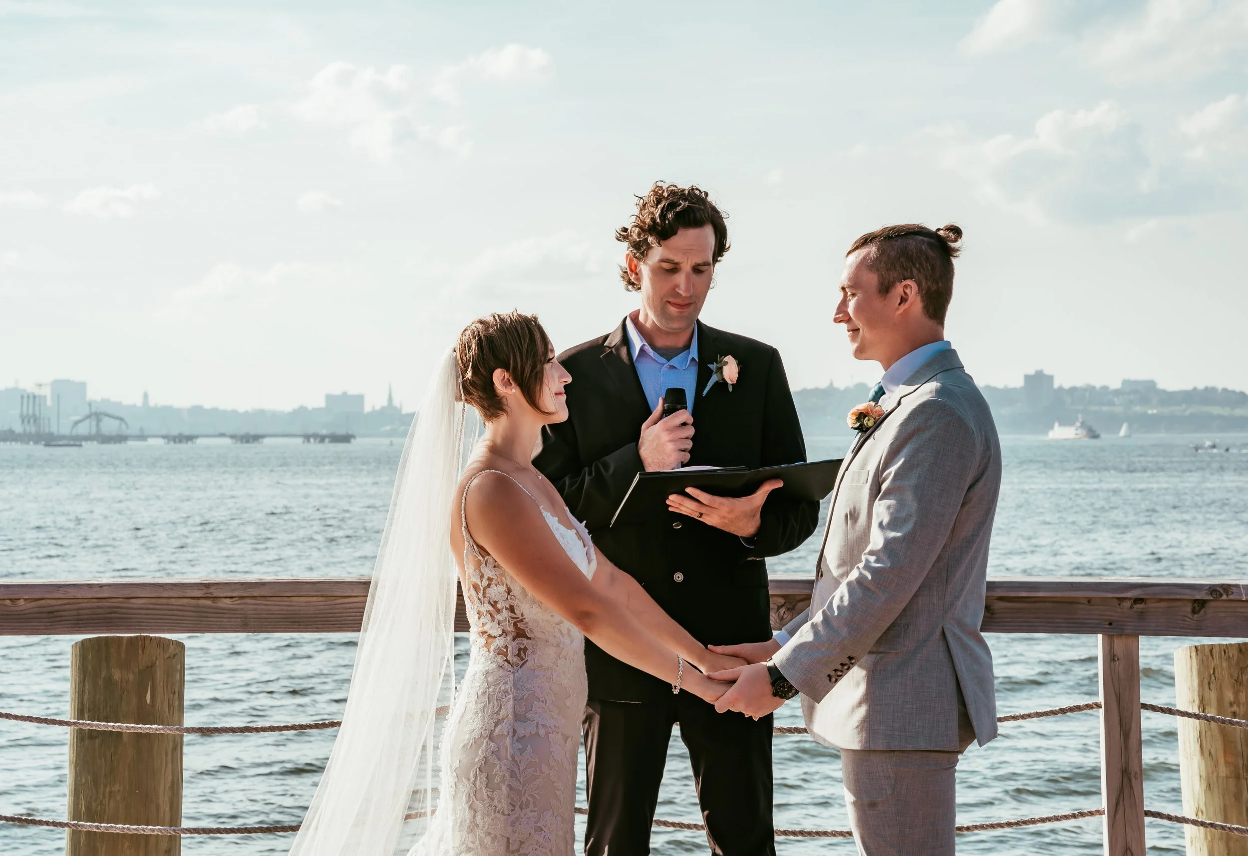A couple getting married outdoors by the water, holding hands and facing each other, with a person officiating ceremony in front of them holding a microphone and reading from a script.