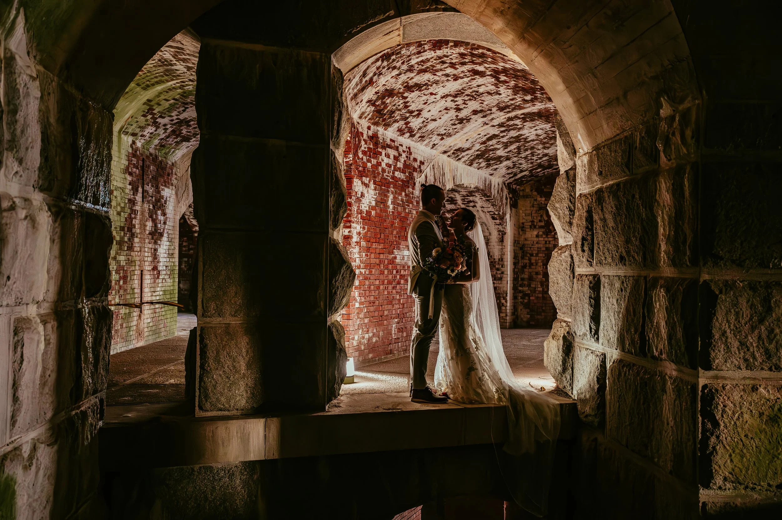 Silhouetted bride and groom standing close together inside a stone and brick archway at their wedding, with the bride holding a bouquet and wearing a veil with a long train.