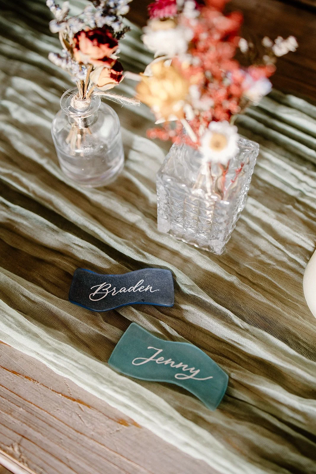 Two small vases with dried flowers on a textured fabric tablecloth, with two fabric name tags reading 'Braden' and 'Jenny'.