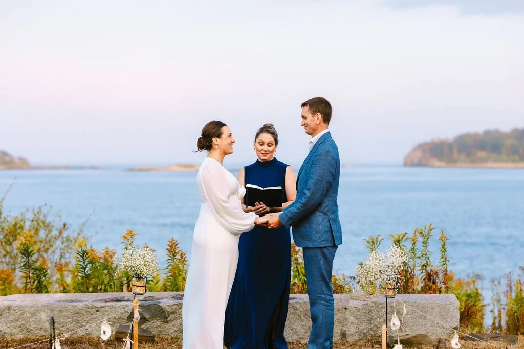 A couple getting married outside by the water with an officiant, holding hands and smiling, during daytime.