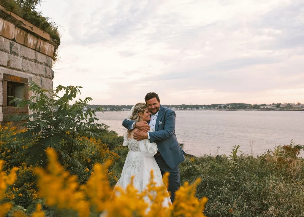 A couple in wedding attire hugging by a river, with greenery and yellow flowers in the foreground, and a cloudy sky.
