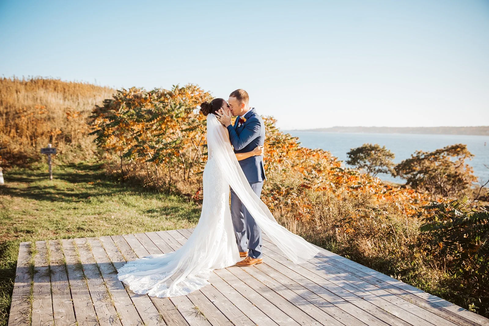 A bride and groom share a kiss on a wooden platform outdoors during their wedding, with autumn-colored trees and lake in the background.