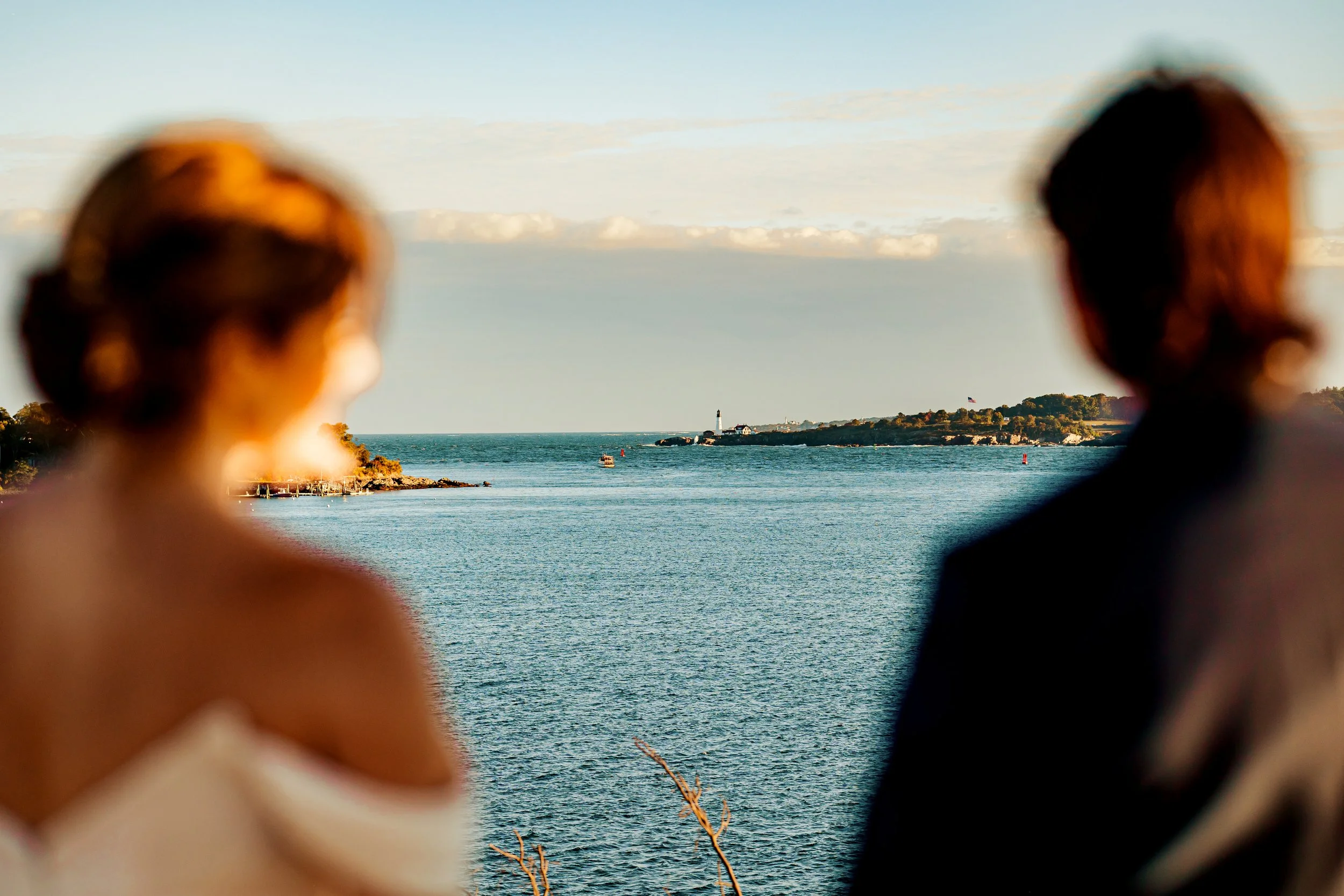 Blurred image of a man and woman facing each other near water, with a lighthouse and coastline visible in the background during golden hour.