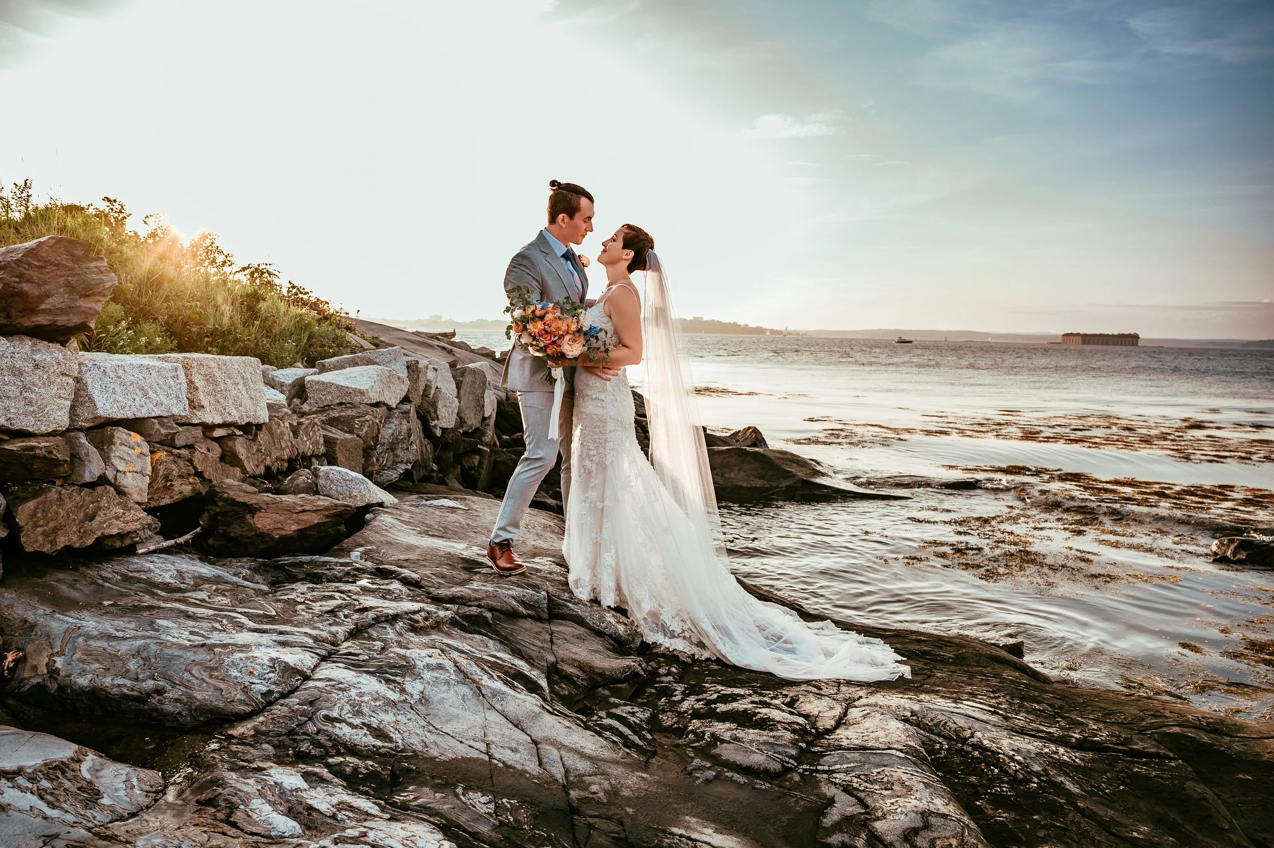 A bride and groom standing on rocks by the water at sunset, with the bride holding a bouquet of flowers and wearing a lace wedding gown, the groom in a gray suit.