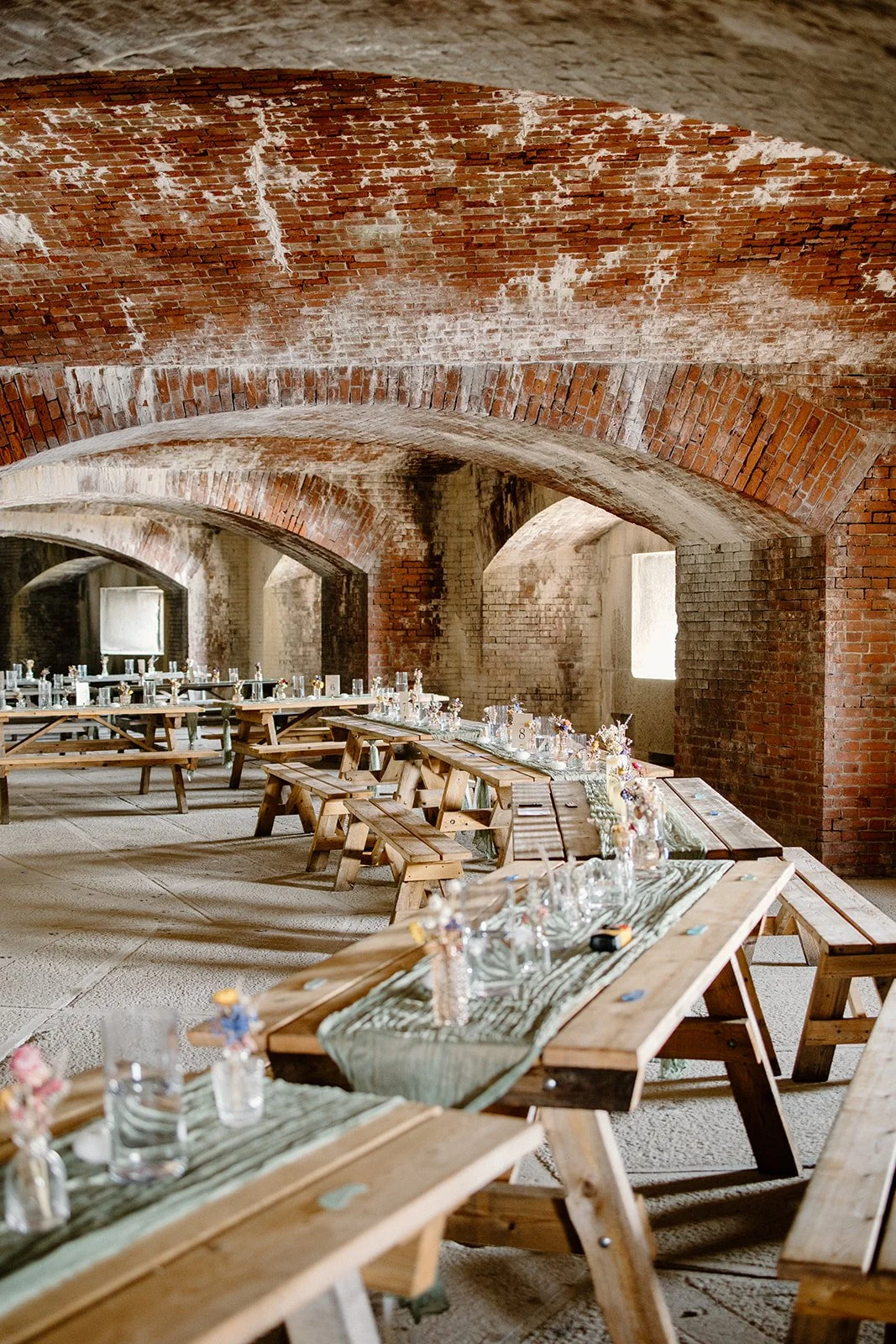 Long wooden tables with small flower arrangements and glassware inside a rustic stone and brick building with arched ceilings and small windows.