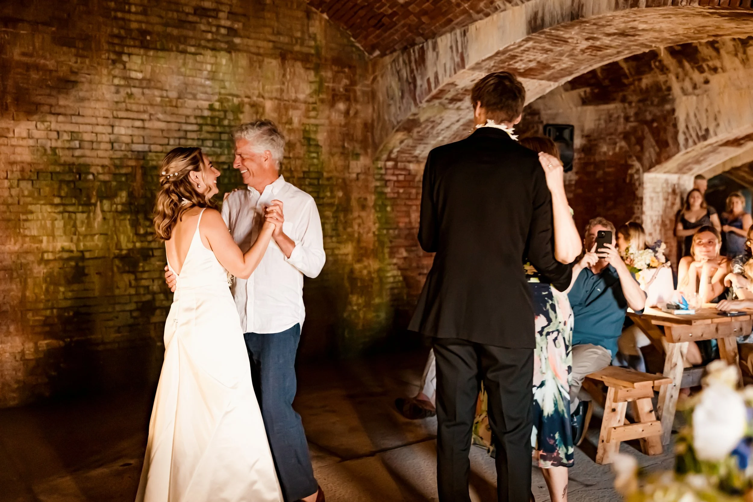 A bride and an older man dancing together at a wedding reception, with guests watching and taking photos in a rustic brick venue.