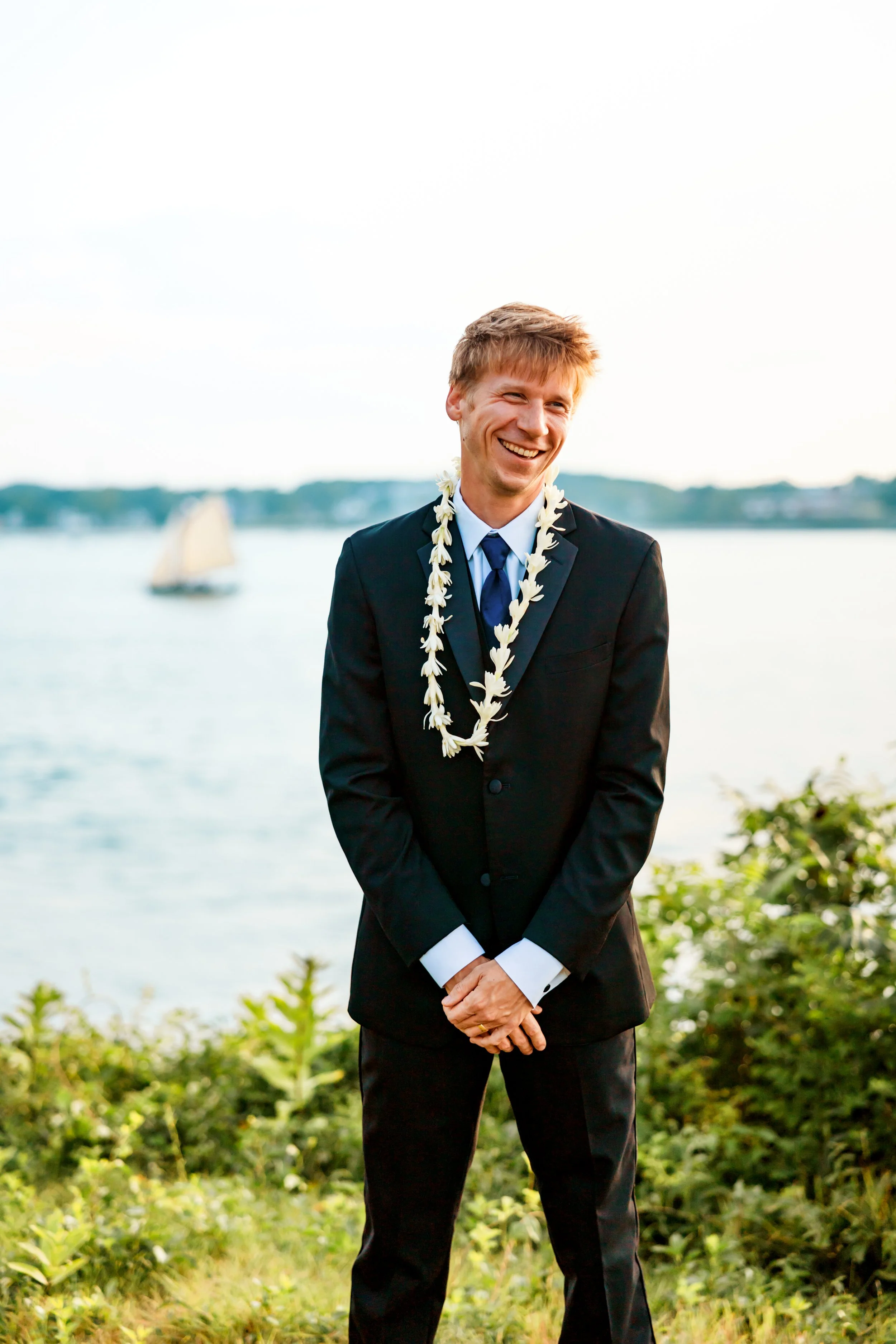 Young man in a black suit, white shirt, and blue tie standing outdoors near water, smiling, wearing a white floral lei.