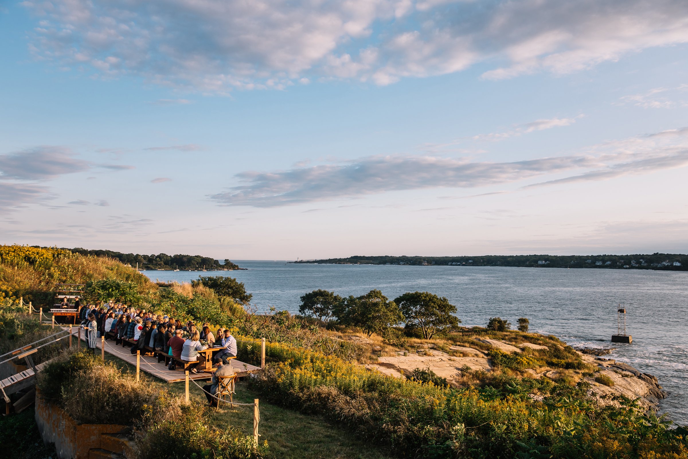 Large group of people sitting at picnic tables outdoors on a hillside overlooking a body of water, with trees and a small lighthouse in the distance under a partly cloudy sky.