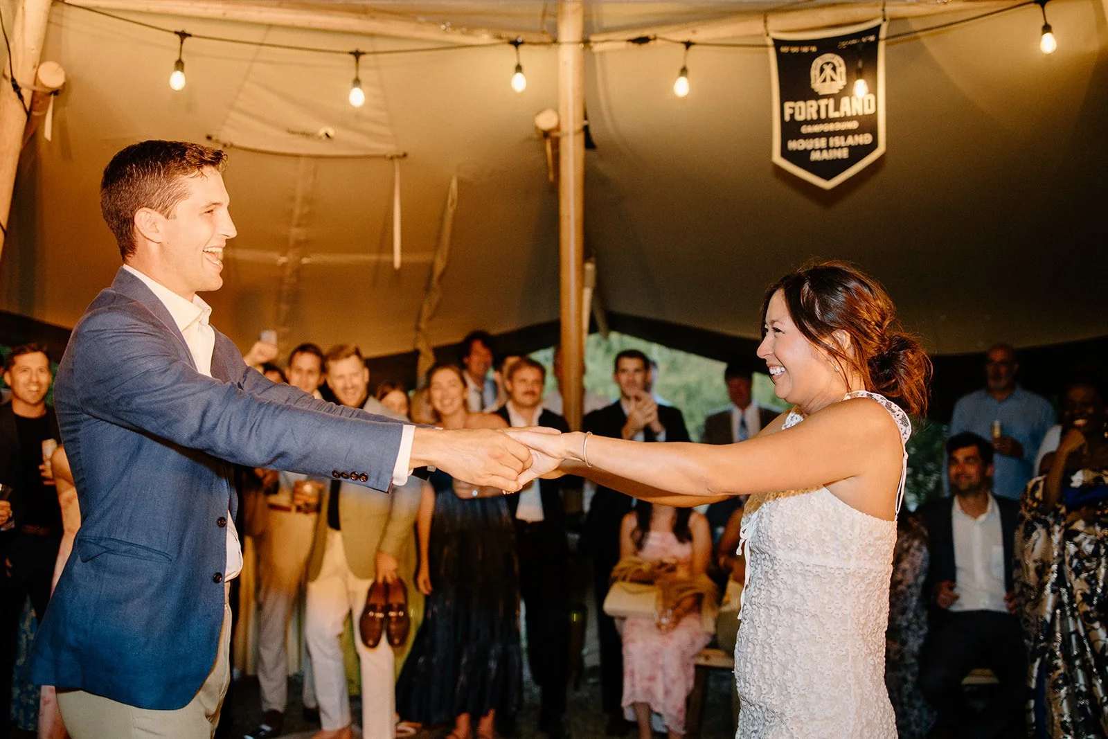 A bride and groom are holding hands and smiling at each other during their wedding reception in a tent with string lights and an audience clapping and watching in the background.