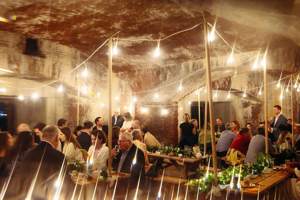 People gathered at a social event or party in a rustic, brick-walled underground space decorated with string lights and greenery.