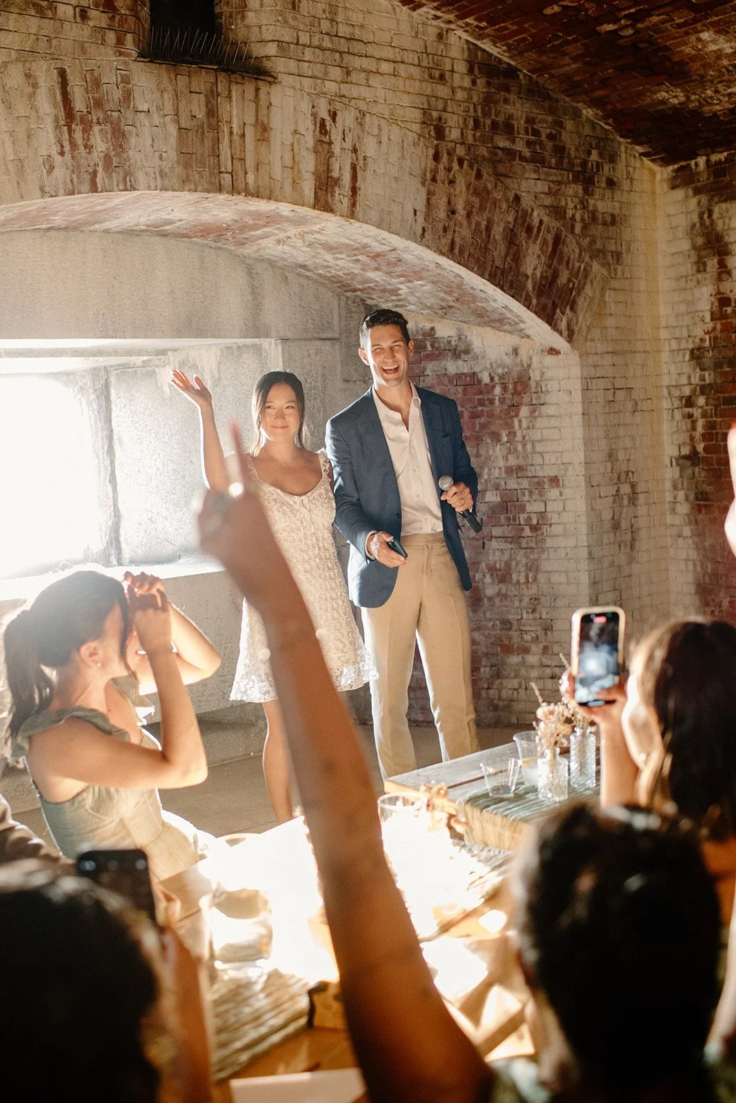 Two people, a man and a woman, stand in front of an audience inside a brick-walled room with exposed arched ceiling, smiling and speaking into microphones.