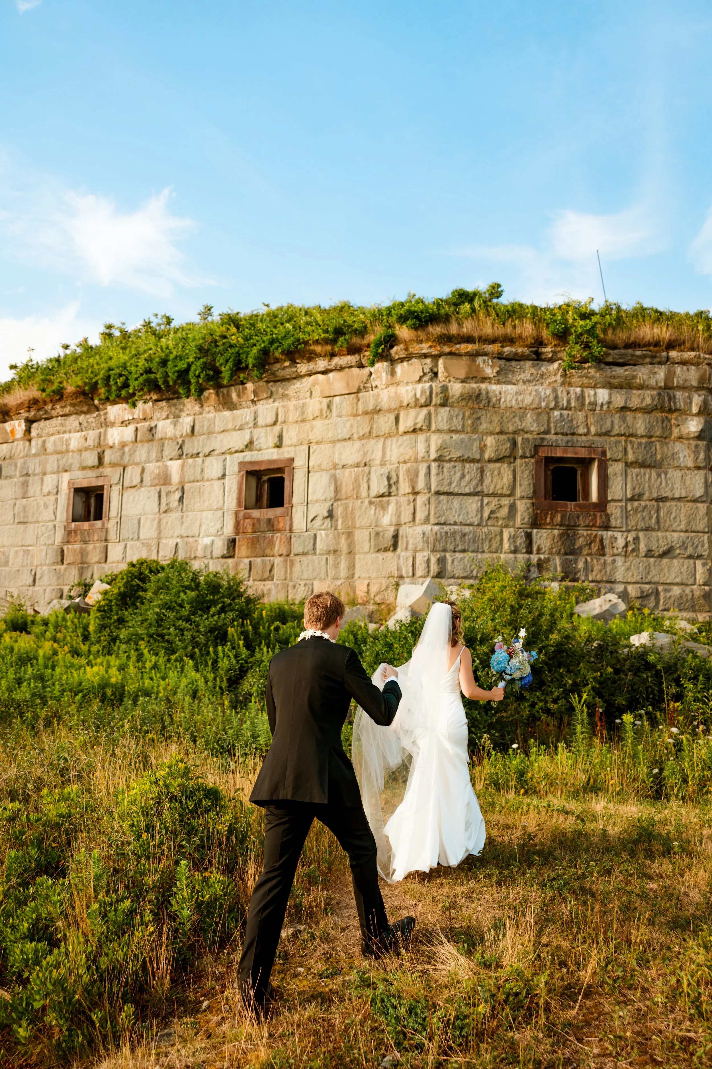 A bride in a white wedding dress and veil holding a bouquet of blue flowers, walking towards an old stone building with greenery, while a groom in a black suit follows her.