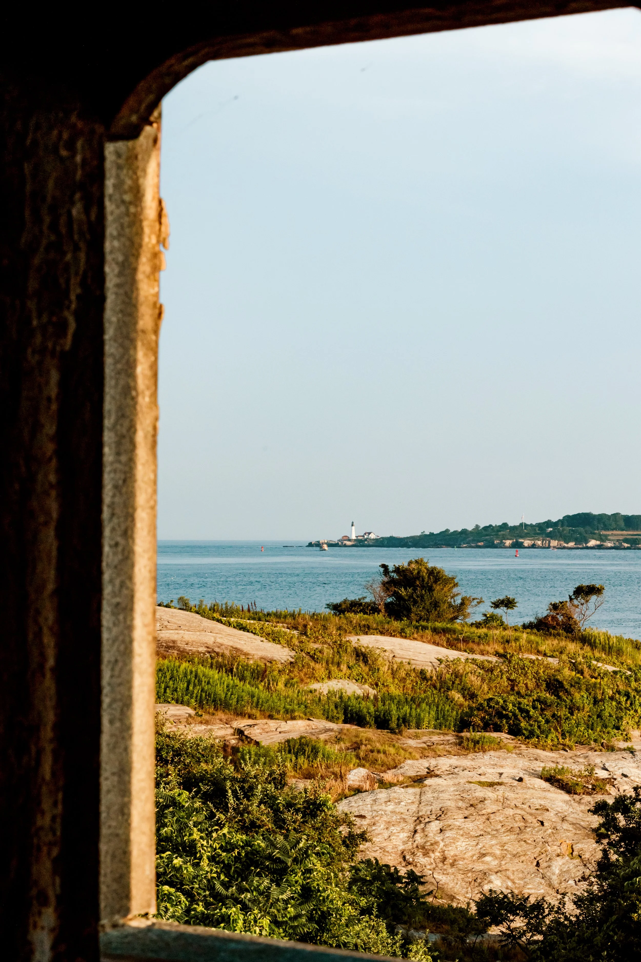 View of a rocky shoreline, greenery, and water with a lighthouse on the distant headland, seen through an arched window or opening.