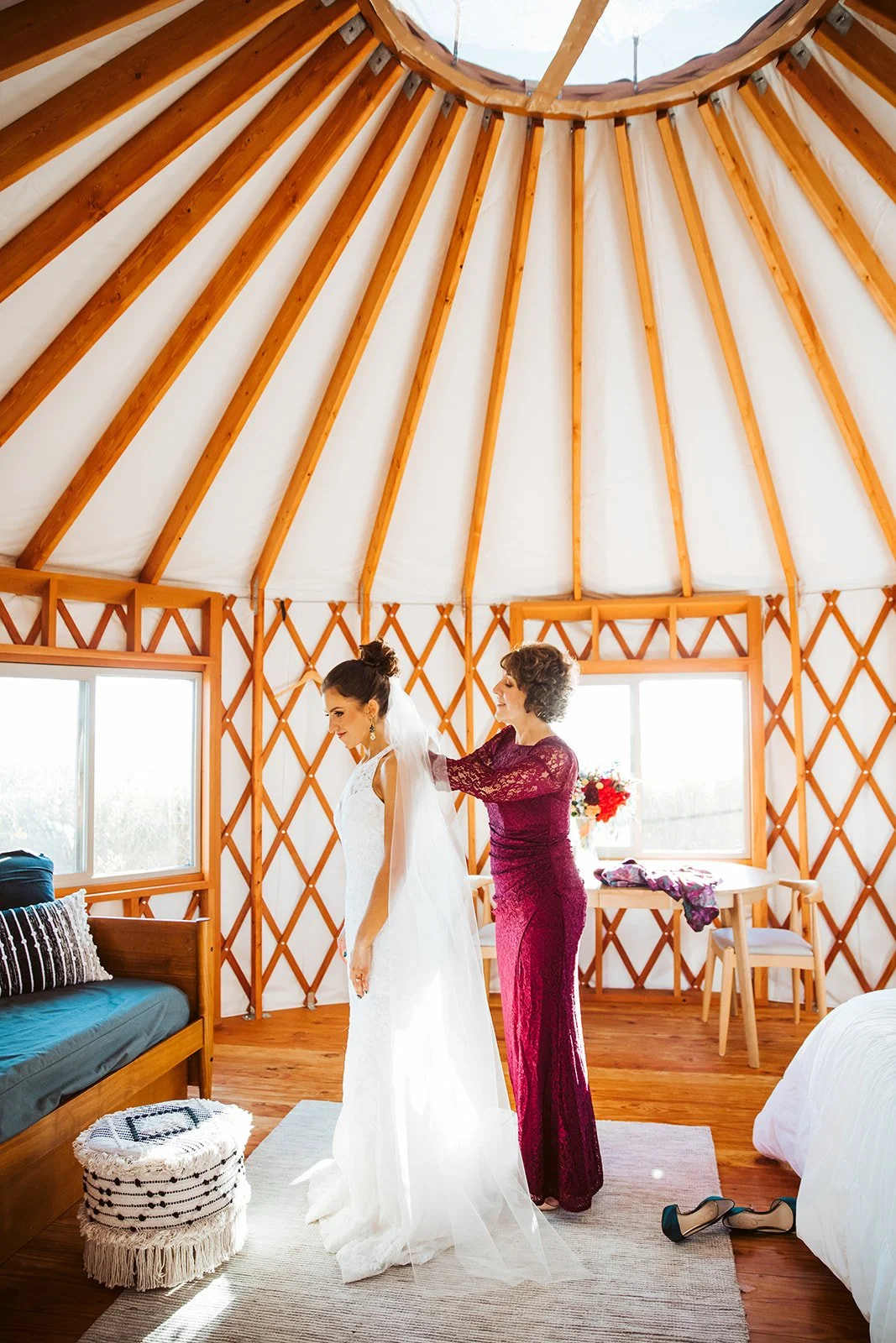A bride in a white wedding dress and veil is being helped with her dress by a woman in a burgundy lace dress inside a yurt with wooden interior and large windows.