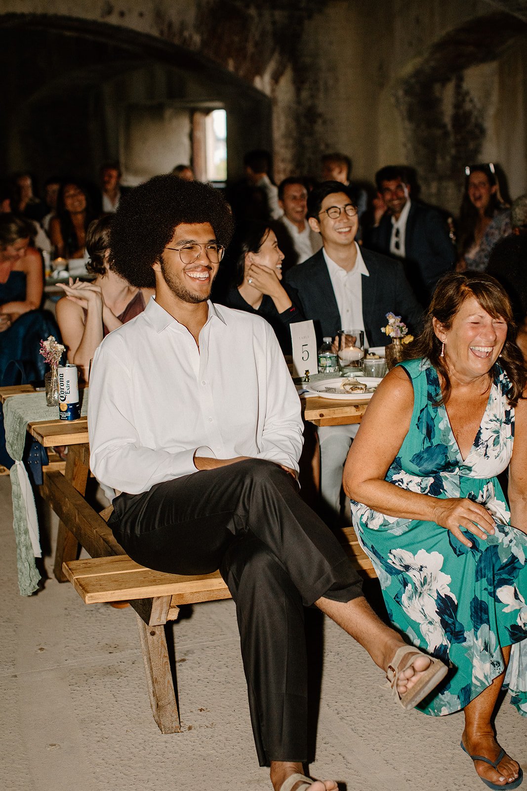 People sitting and laughing at a wedding or social gathering in a rustic venue, some with drinks and table decorations.