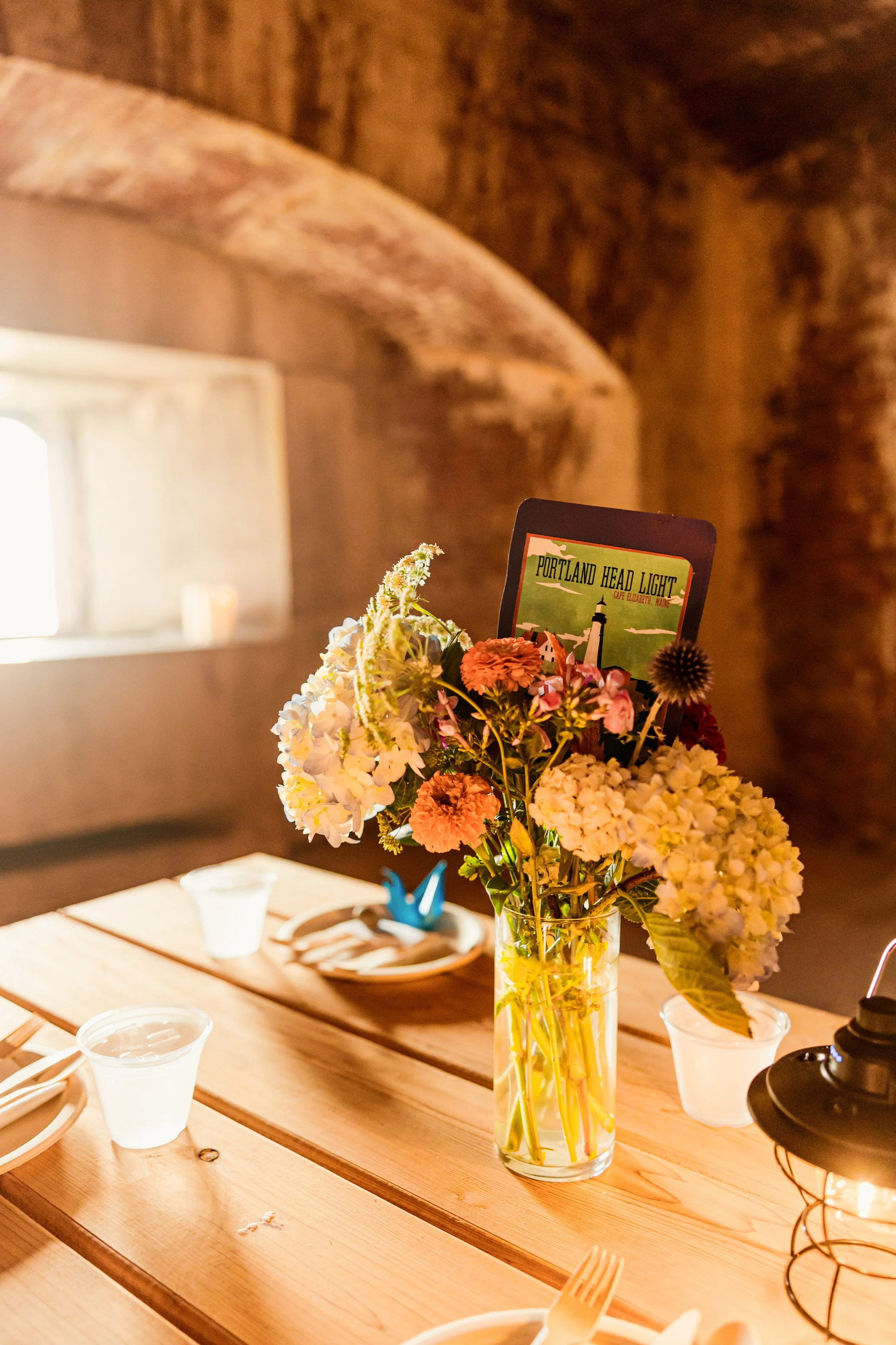 A vase of mixed colorful flowers on a wooden table in a cozy, rustic room with a brick wall and a window.