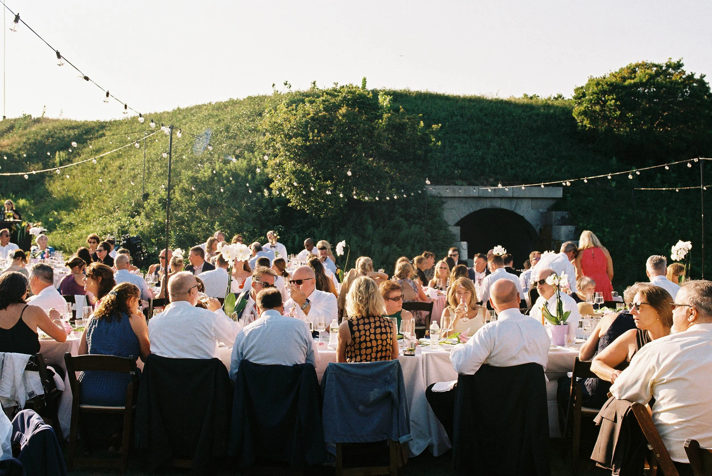 Outdoor banquet with many guests seated at round tables, decorated with white orchids and tableware, under string lights, with greenery and a tunnel in the background.
