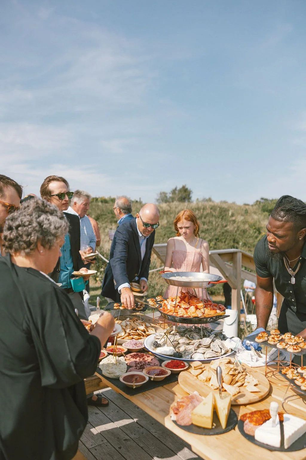 People gathered outdoors around a table with various foods including seafood, cheese, meats, and appetizers, enjoying a meal on a sunny day.