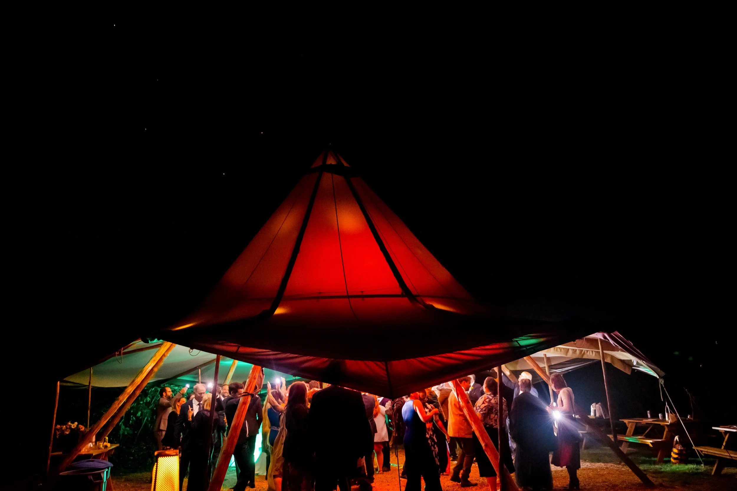 People dancing at a nighttime outdoor event under a large illuminated tent with string lights and picnic tables nearby.