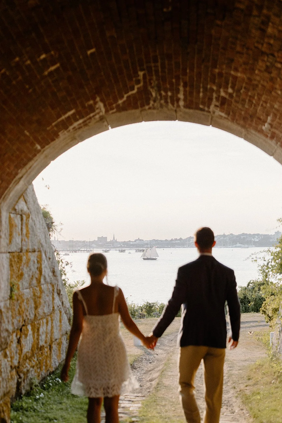 A couple holding hands, walking under a brick tunnel toward a body of water with sailboats and a distant city skyline.