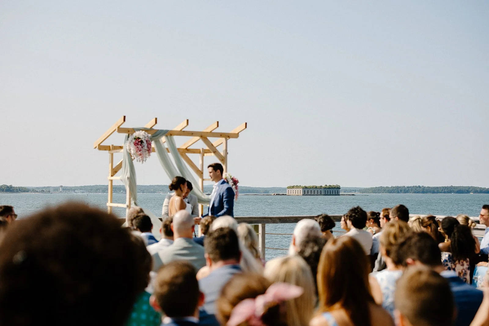 A wedding ceremony taking place outdoors by the water, with a wooden arch decorated with flowers and fabric, and a bride and groom standing under it, surrounded by guests.