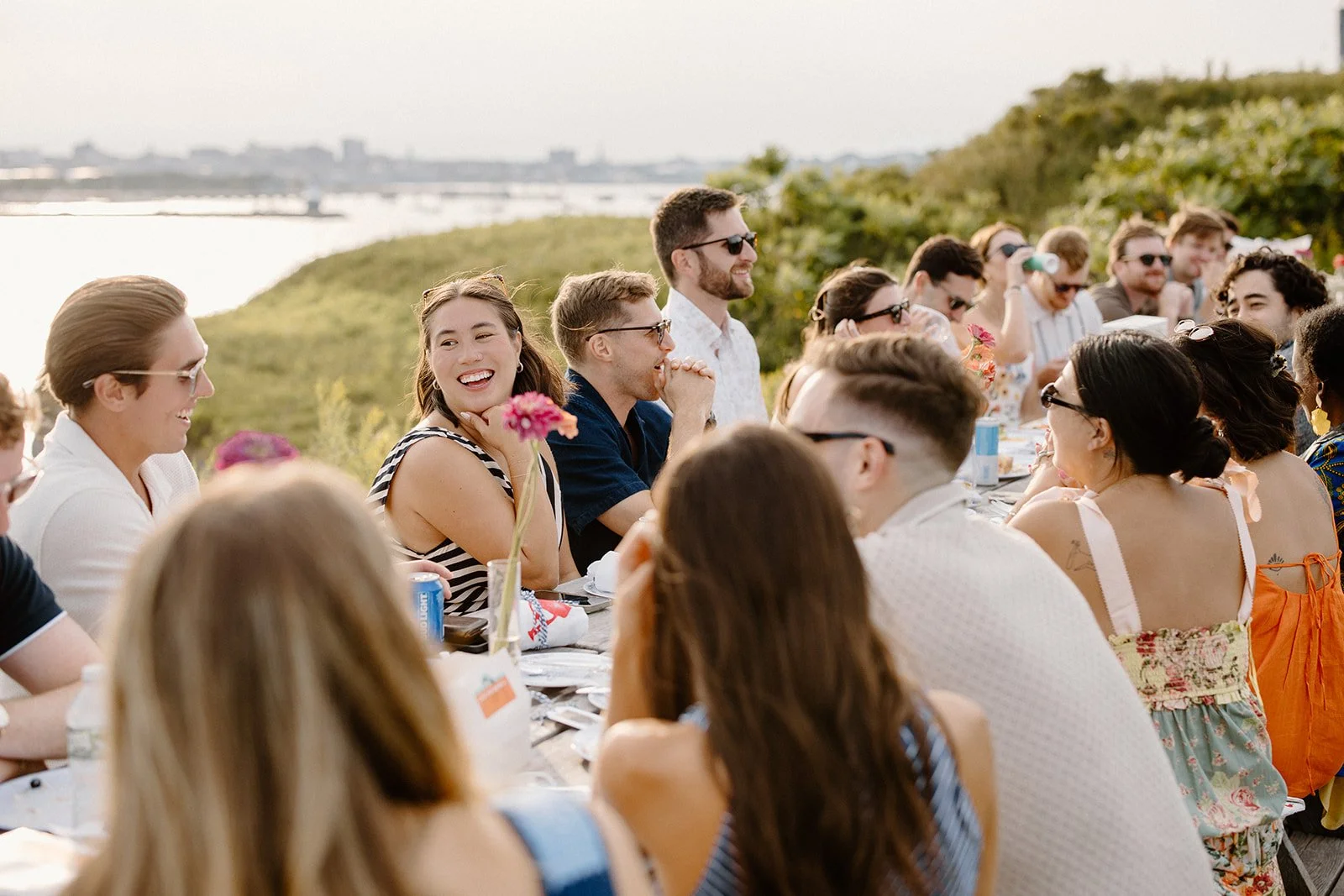 A group of people gathered around a long outdoor table in a scenic setting with water and greenery in the background, enjoying a meal and conversation.