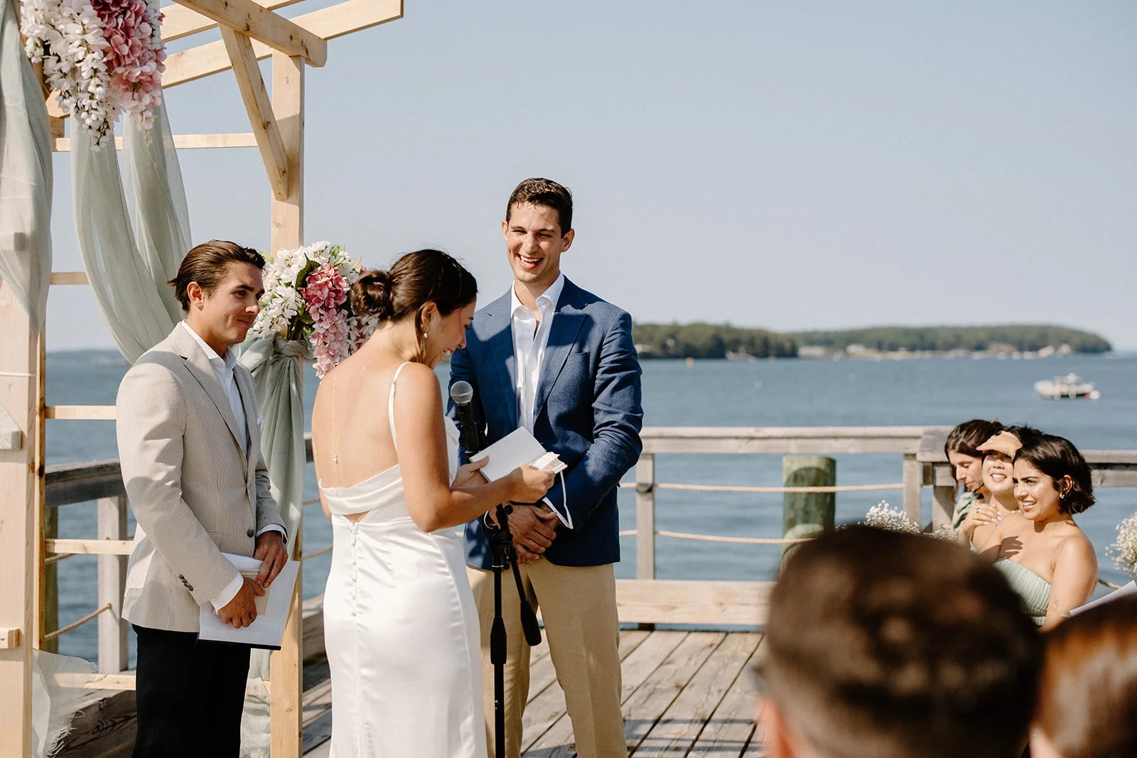 A couple gets married outdoors by the water, with a officiant reading vows and guests watching on, on a wooden deck with floral decorations.
