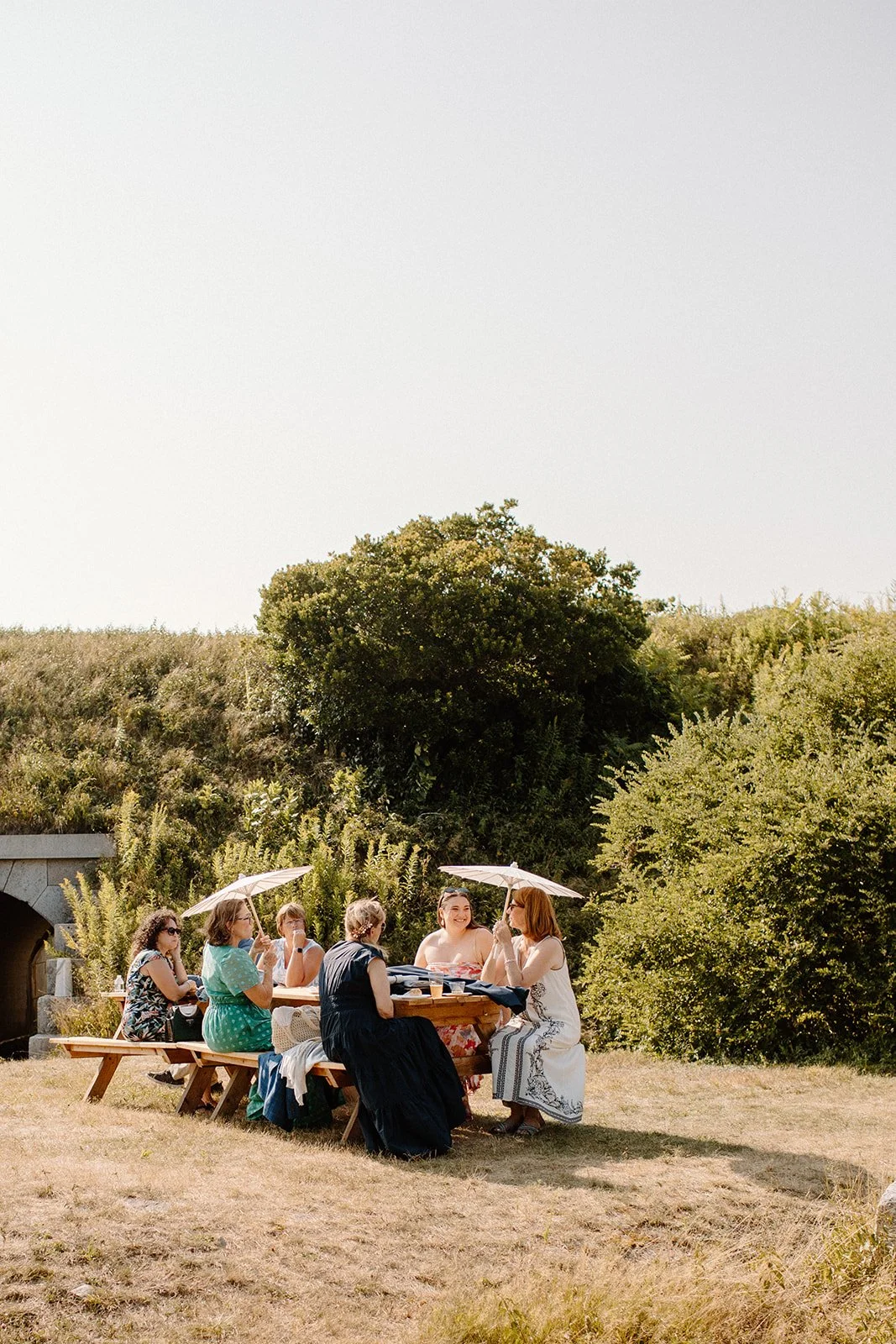 A group of women sitting around a wooden picnic table outdoors, some holding umbrellas, enjoying a sunny day with green trees and bushes in the background.