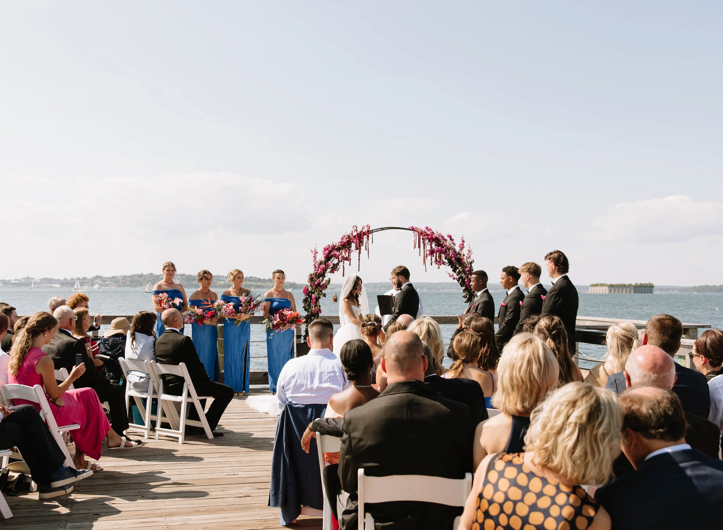 A seaside wedding ceremony with a floral arch, officiant, bridesmaids in blue dresses holding flowers, and groomsmen in dark suits, with guests seated on a wooden deck near the water under a partly cloudy sky.