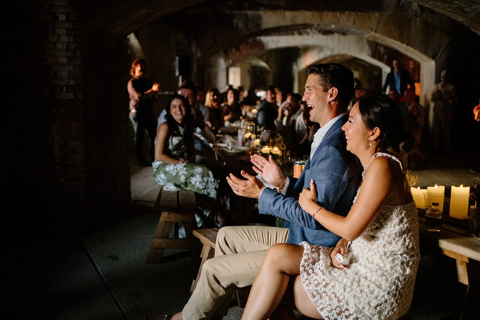 A wedding reception with guests sitting at a long table, two people in the foreground are laughing and clapping, surrounded by candles and dim lighting.