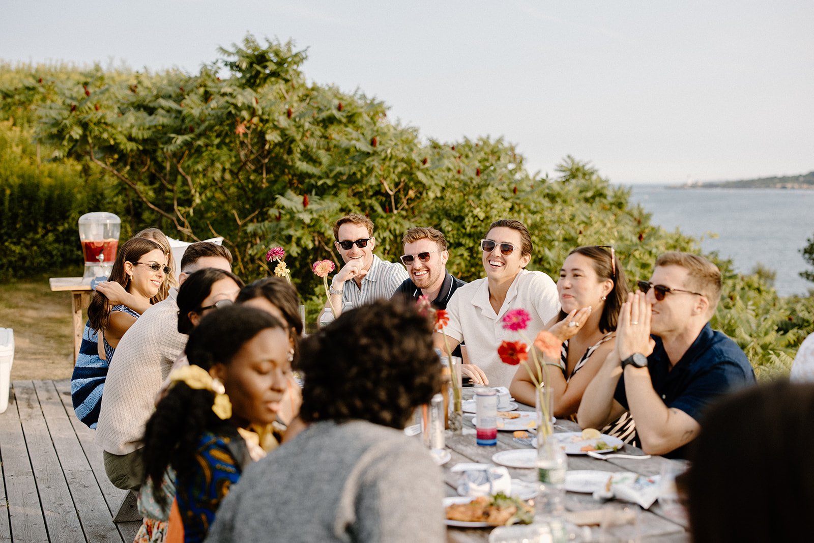 A group of people enjoying a meal outdoors at a long table near water, with greenery and trees in the background, on a sunny day.