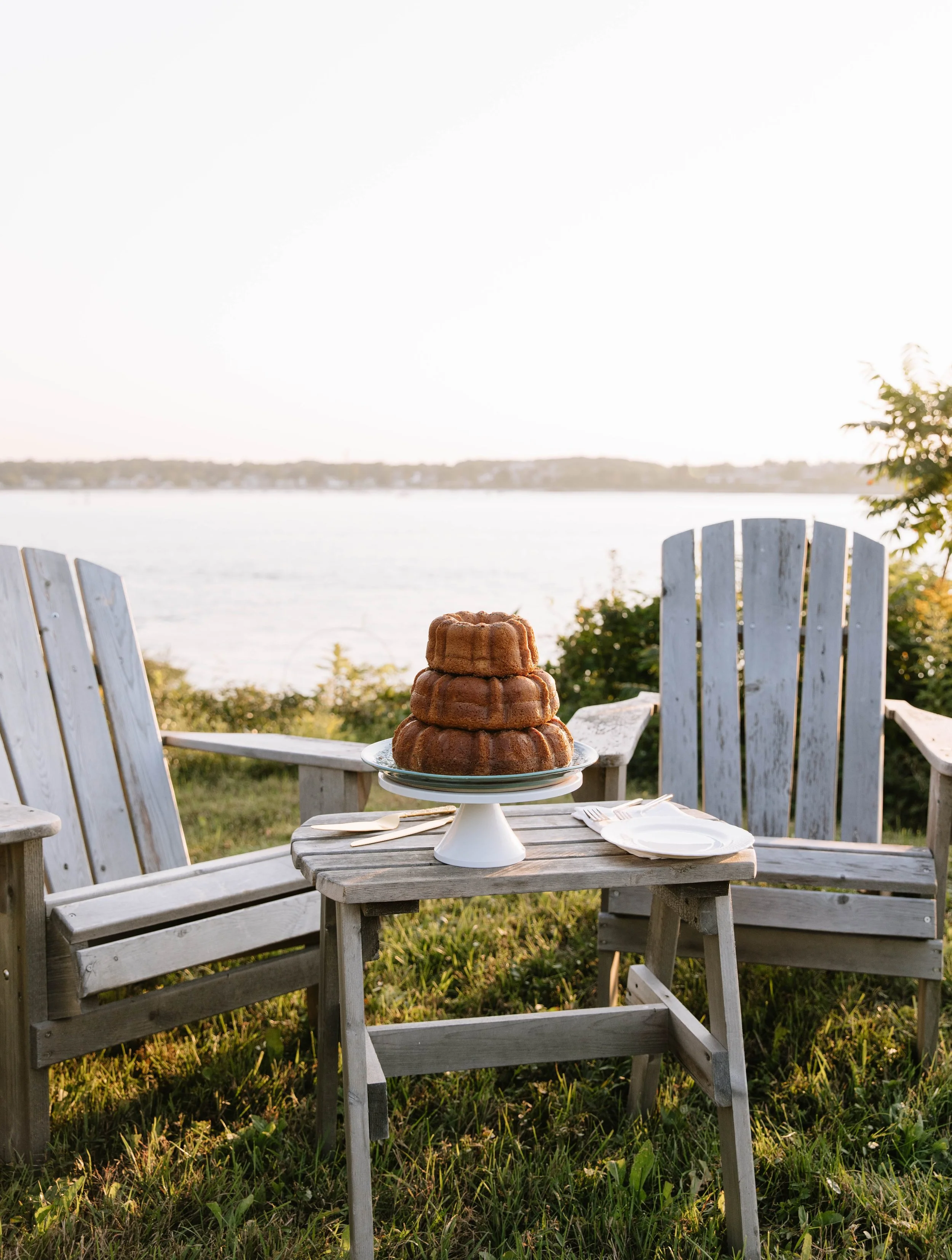 A Bundt cake on a cake stand placed on a small wooden table between two Adirondack chairs, outdoors near a body of water, during sunset.