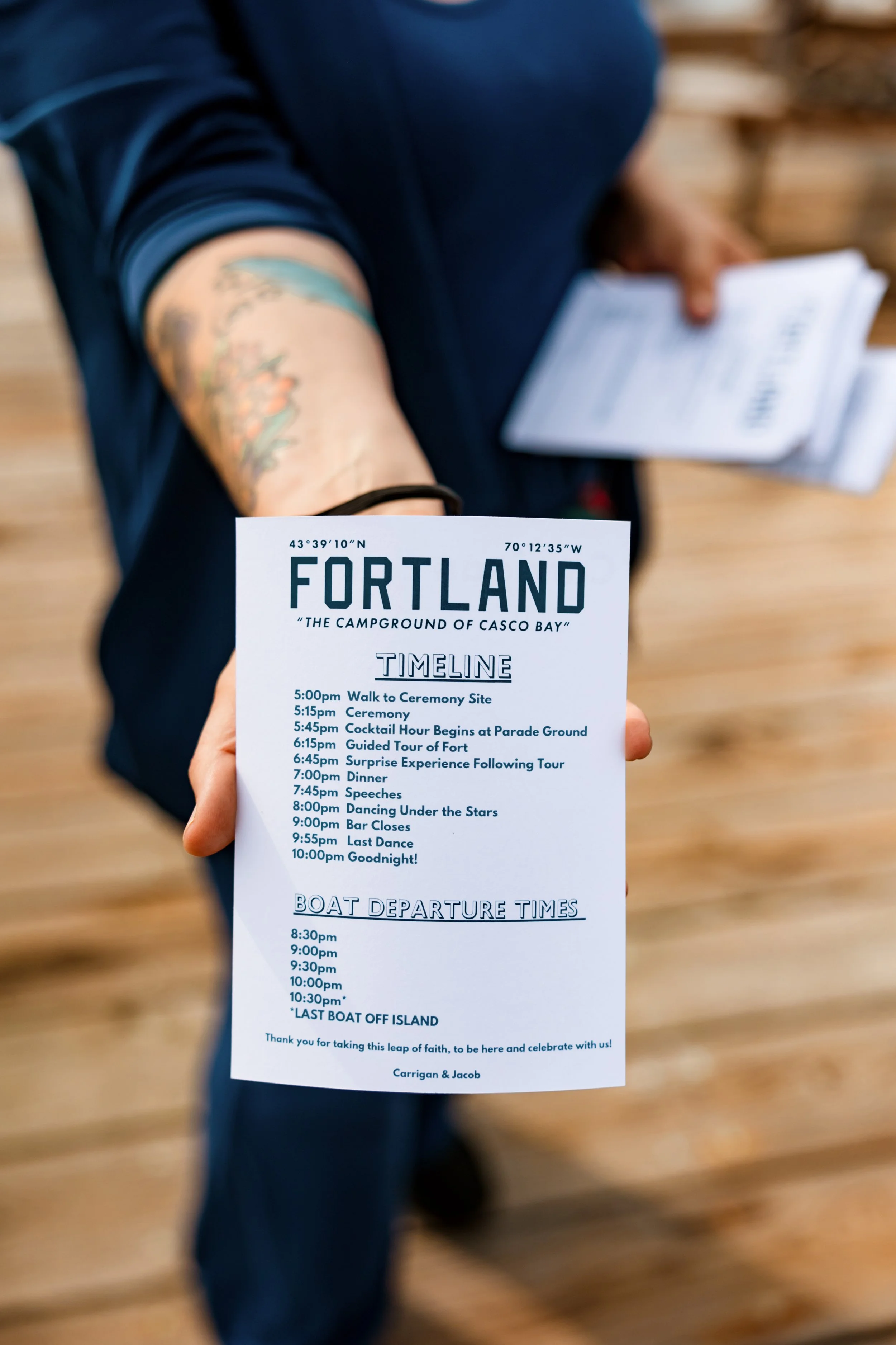A person holding a detailed event schedule for Fortland, Casco Bay. The schedule includes times for activities like walking to the ceremony site, the ceremony, cocktails, a tour of the fort, dinner, speeches, dancing, bar closing, and boat departure 