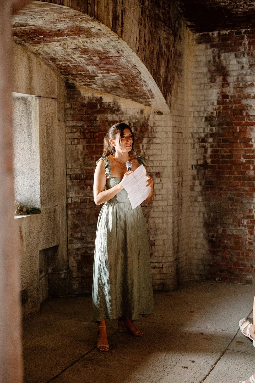 A woman in a green dress holding a microphone and paper, speaking at an event in an indoor setting with exposed brick walls and natural light.