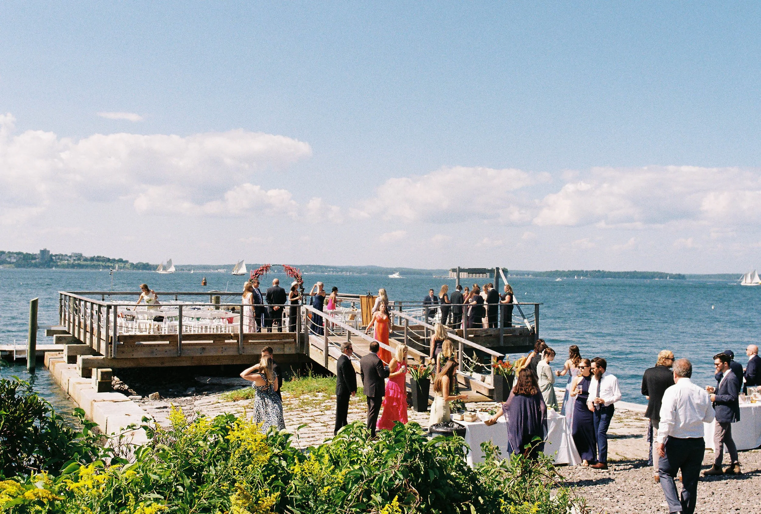 People celebrating at an outdoor waterfront event on a bright, sunny day with sailboats on the water.