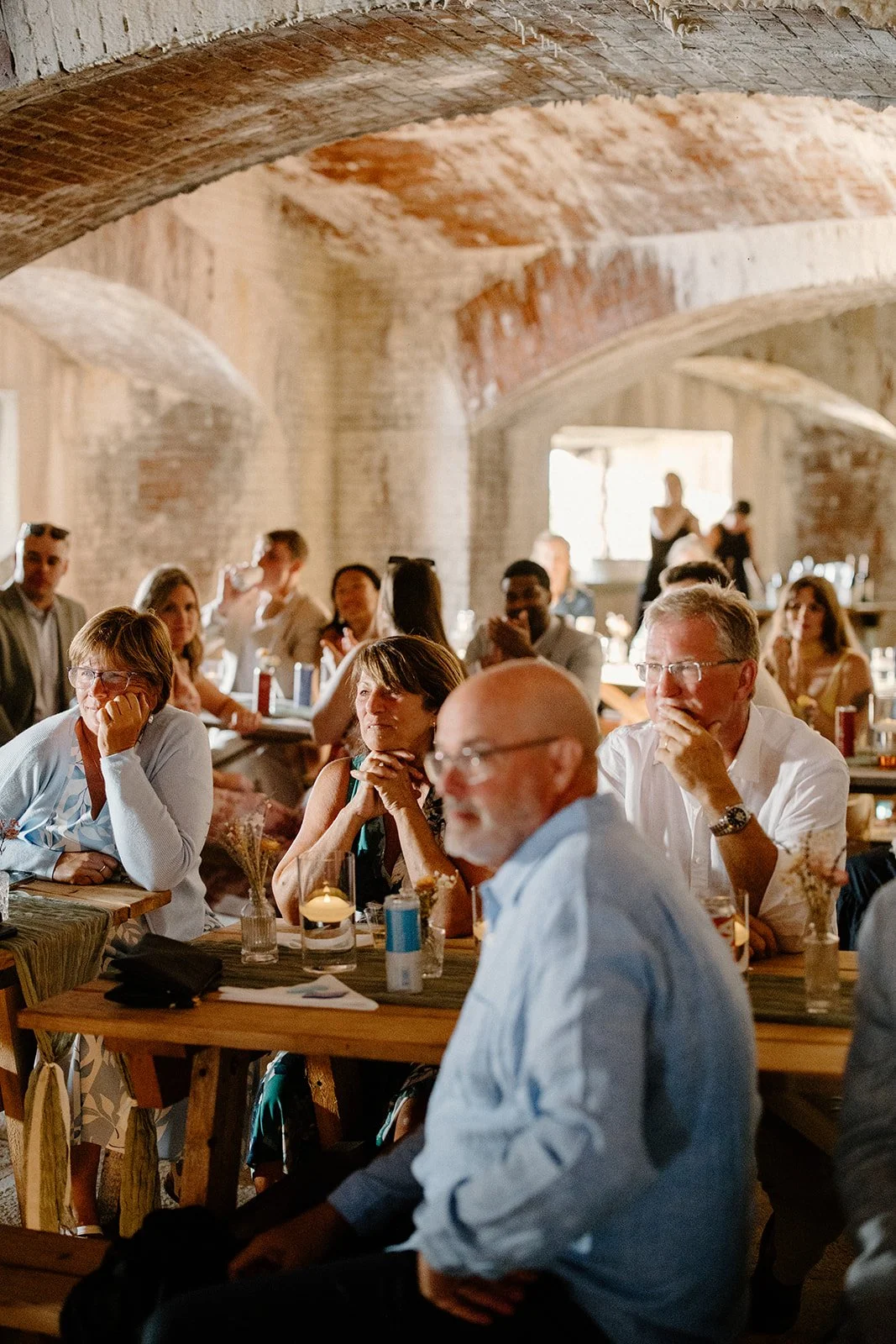 People sitting at wooden tables in a rustic brick-walled venue, attentively listening, with some glasses, drinks, and flower arrangements on the tables.